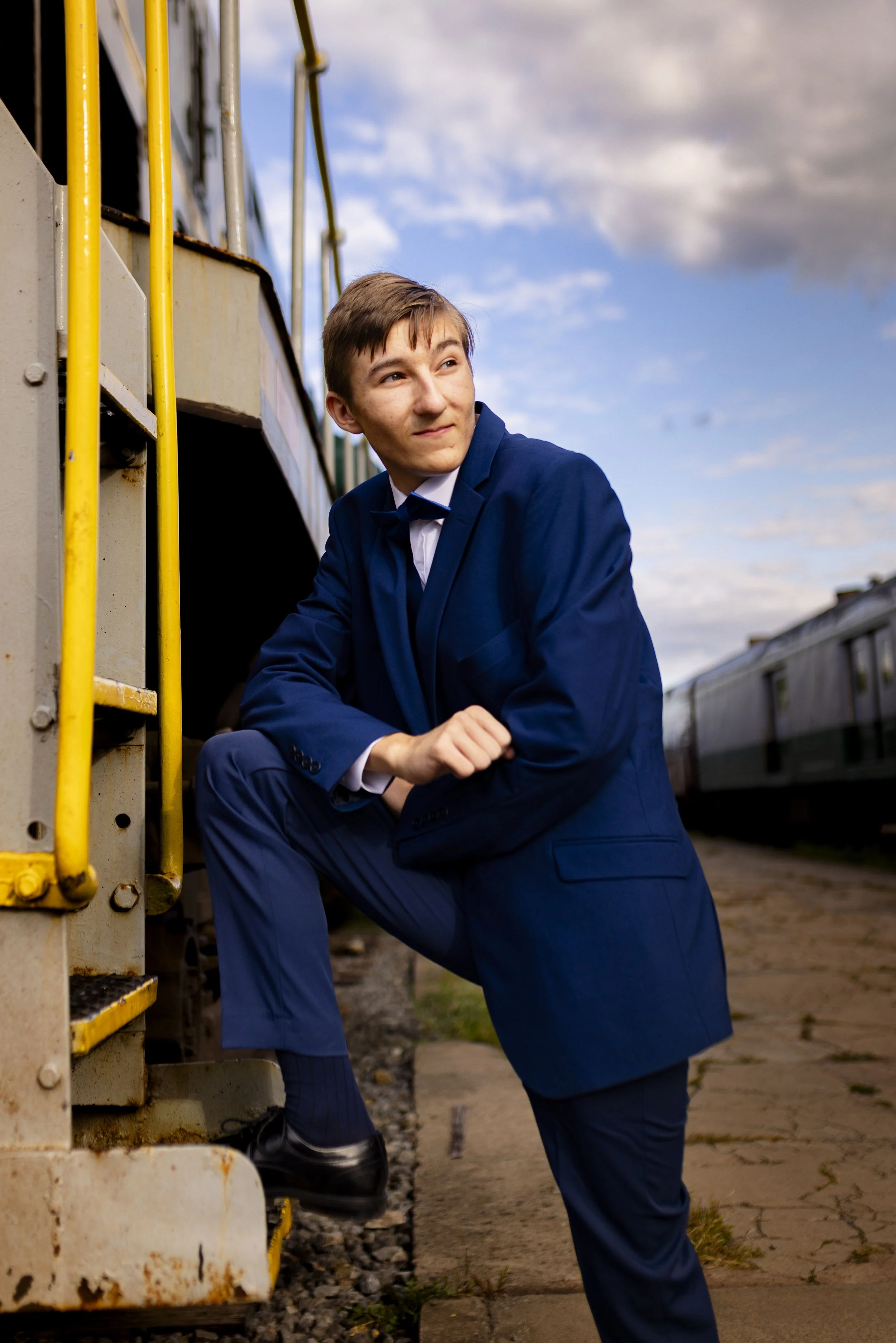 A young man in a navy blue suit kneels next to a train, looking to the side with a slight smile, on a train platform during daytime.
