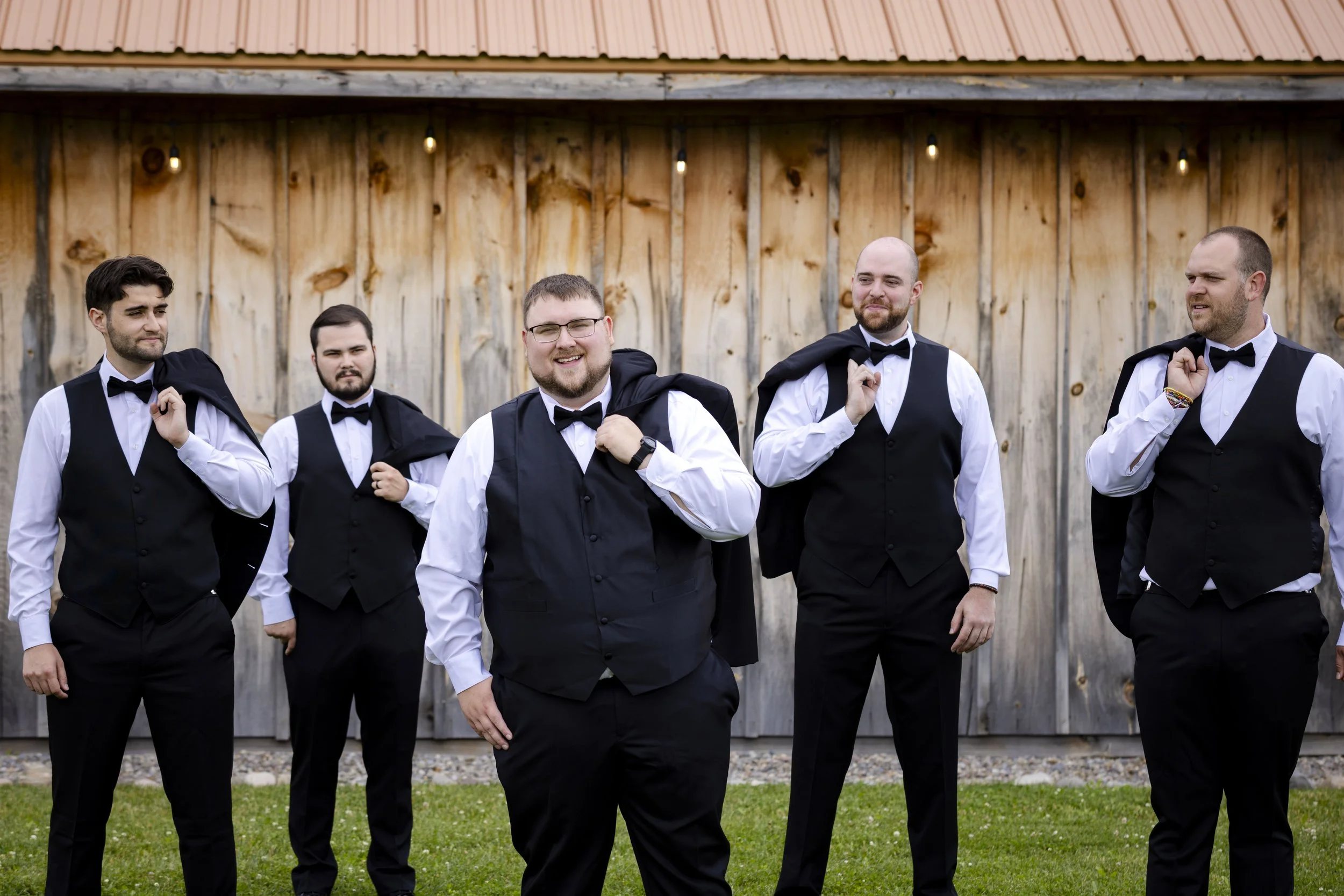 Group of five men dressed in tuxedos with black vests, white shirts, and black bow ties, standing outdoors in front of a wooden fence, with some holding jackets over their shoulders.