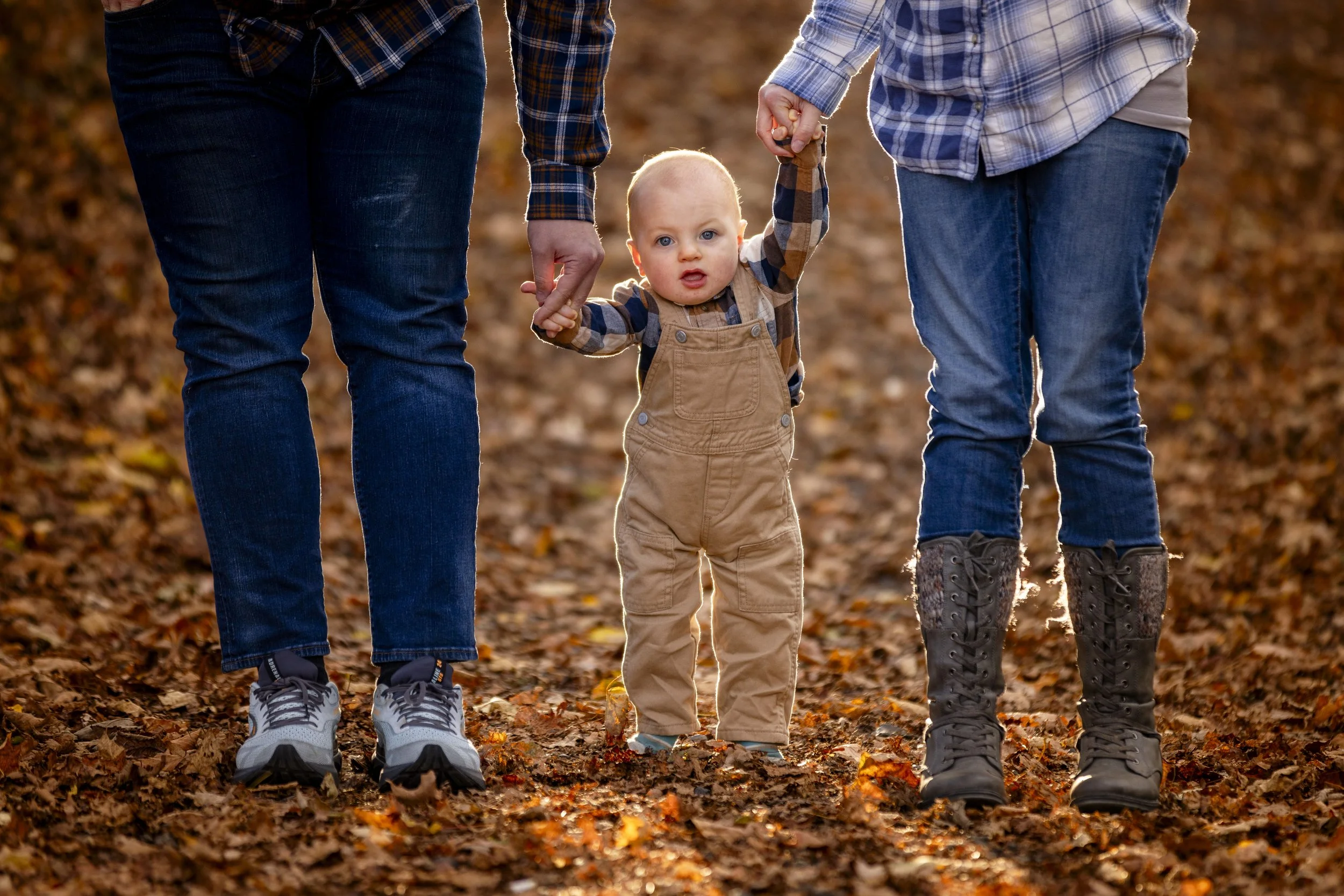 A toddler holding hands with two adults, walking on a path covered with autumn leaves.