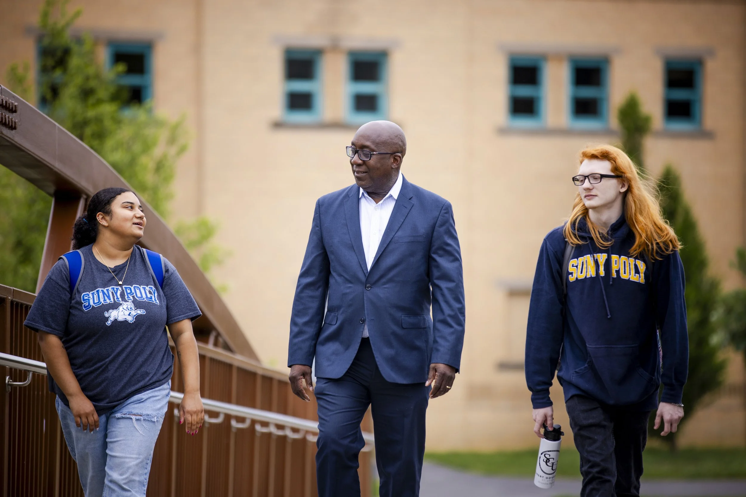 Two students walking with a man in a navy suit and glasses on a university campus with brick buildings and green trees in the background.