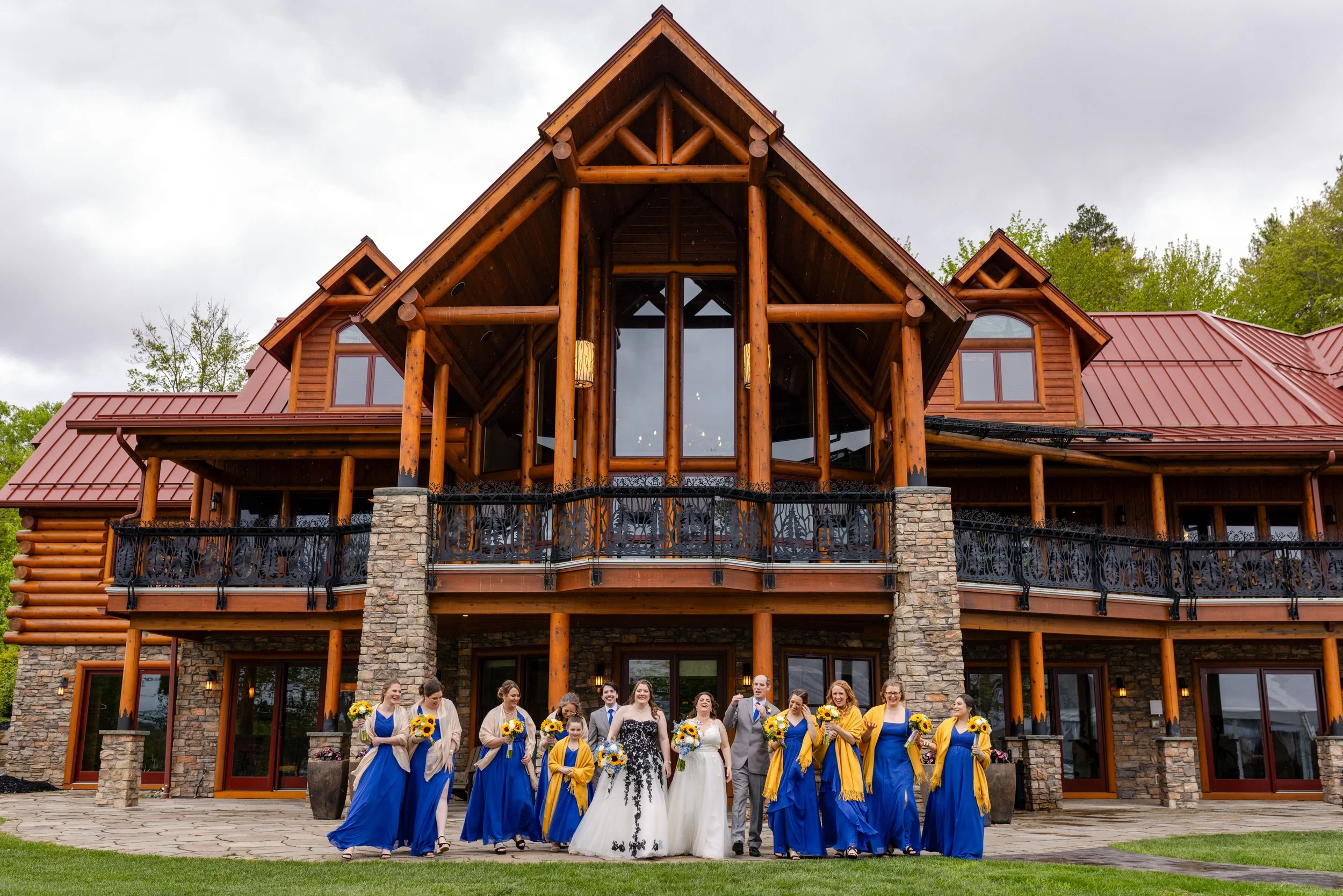Group of people in wedding attire standing in front of a large lodge-style building with wood and stone exterior, patio, and balcony.