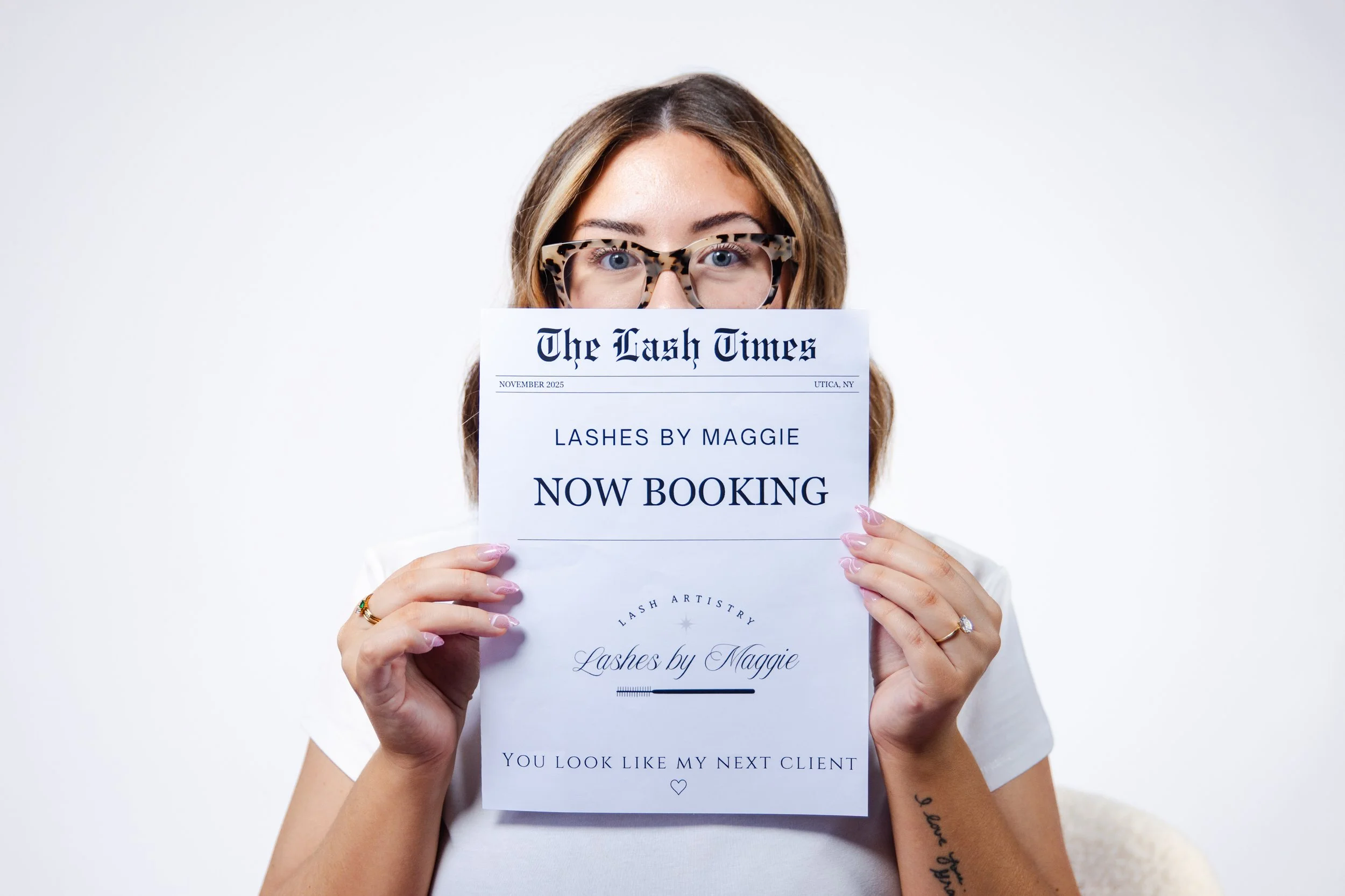 Woman with tortoise-shell glasses holding a paper in front of her face with the headline 'Now Booking' from Lash by Maggie.