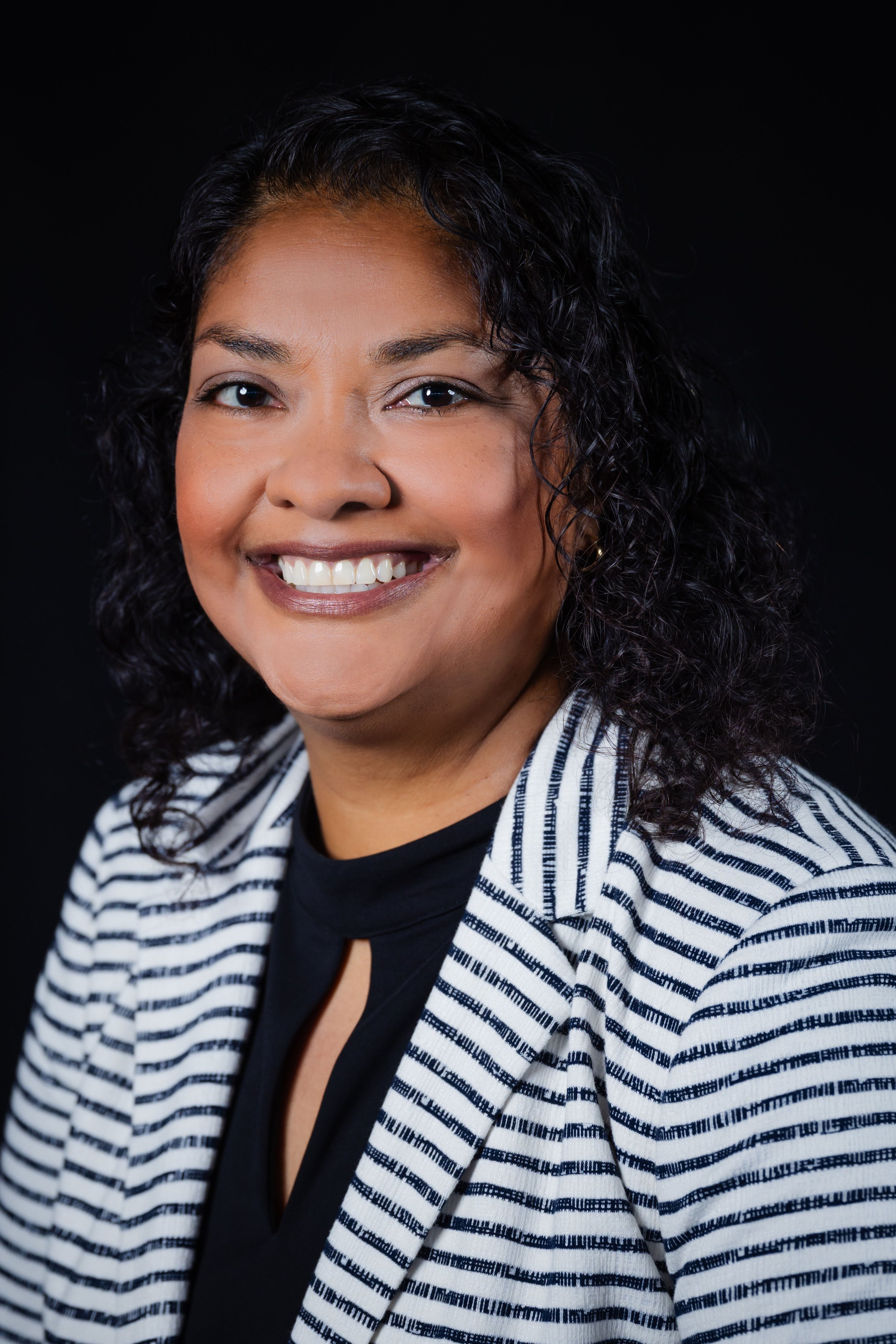 Close-up portrait of a woman with curly black hair and brown eyes, smiling, wearing a black top and a white blazer with black stripes, against a black background.