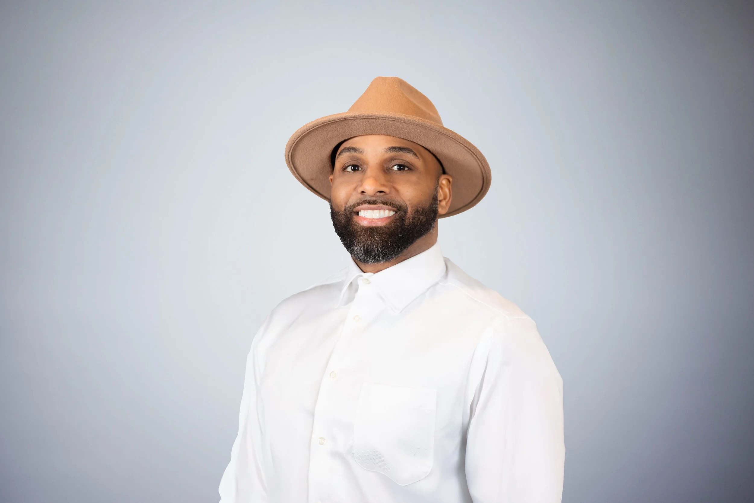 A smiling man with a beard wearing a white button-up shirt and a tan wide-brimmed hat, posing against a light gray background.