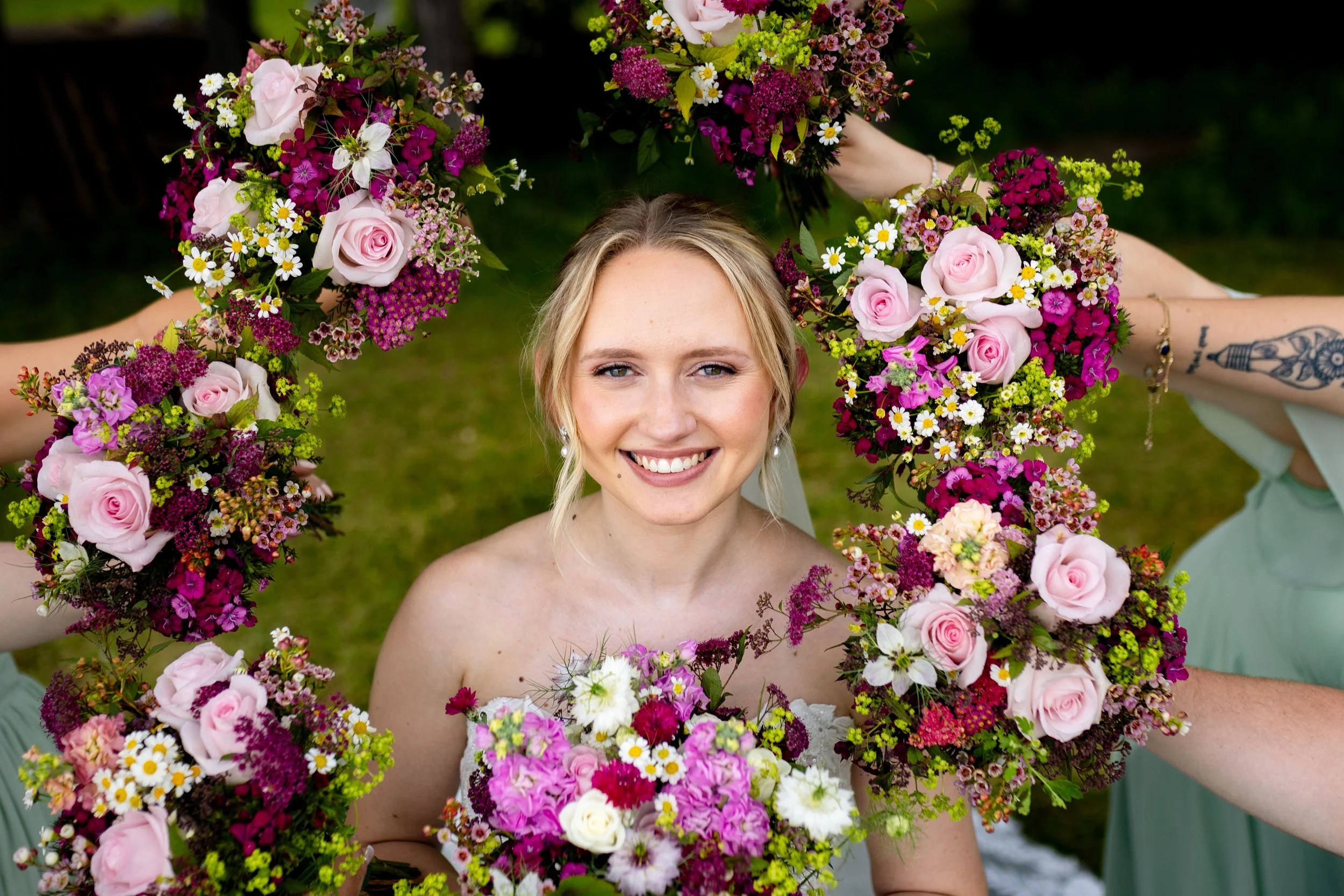A smiling woman with blonde hair and earrings, surrounded by a floral wreath with pink roses, white daisies, and purple flowers, outdoors on a grassy area.