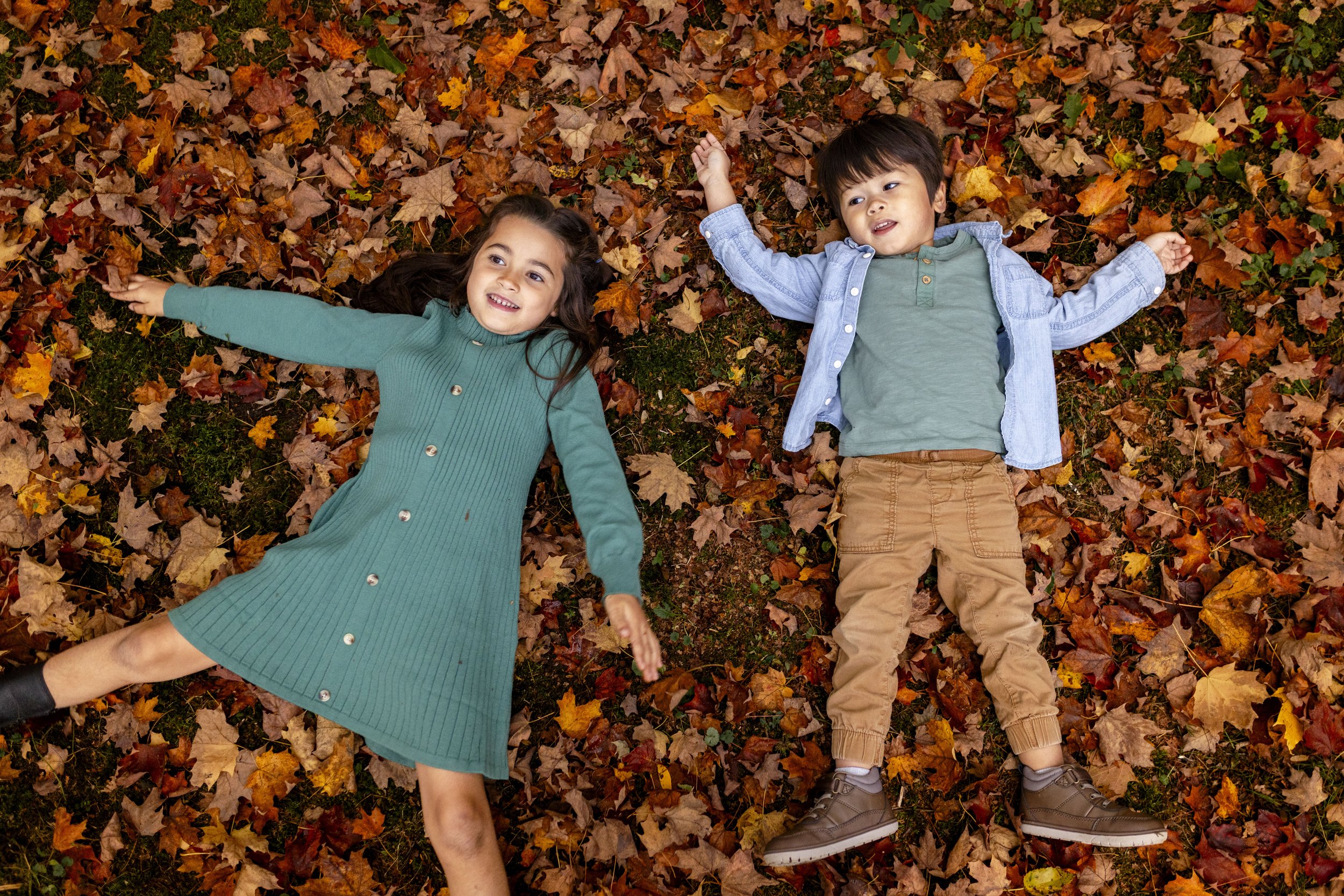 Two children, a girl and a boy, lying on the ground covered with autumn leaves, smiling and looking up.