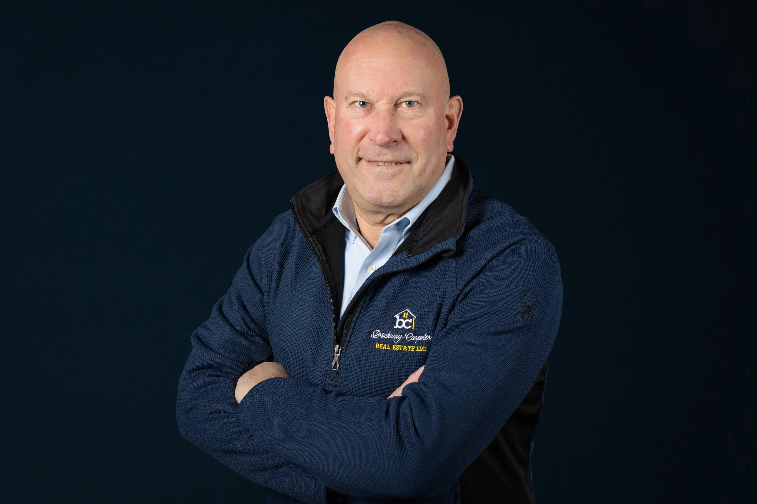 Portrait of a bald man wearing a navy blue jacket with a logo and text, standing with arms crossed against a dark background.