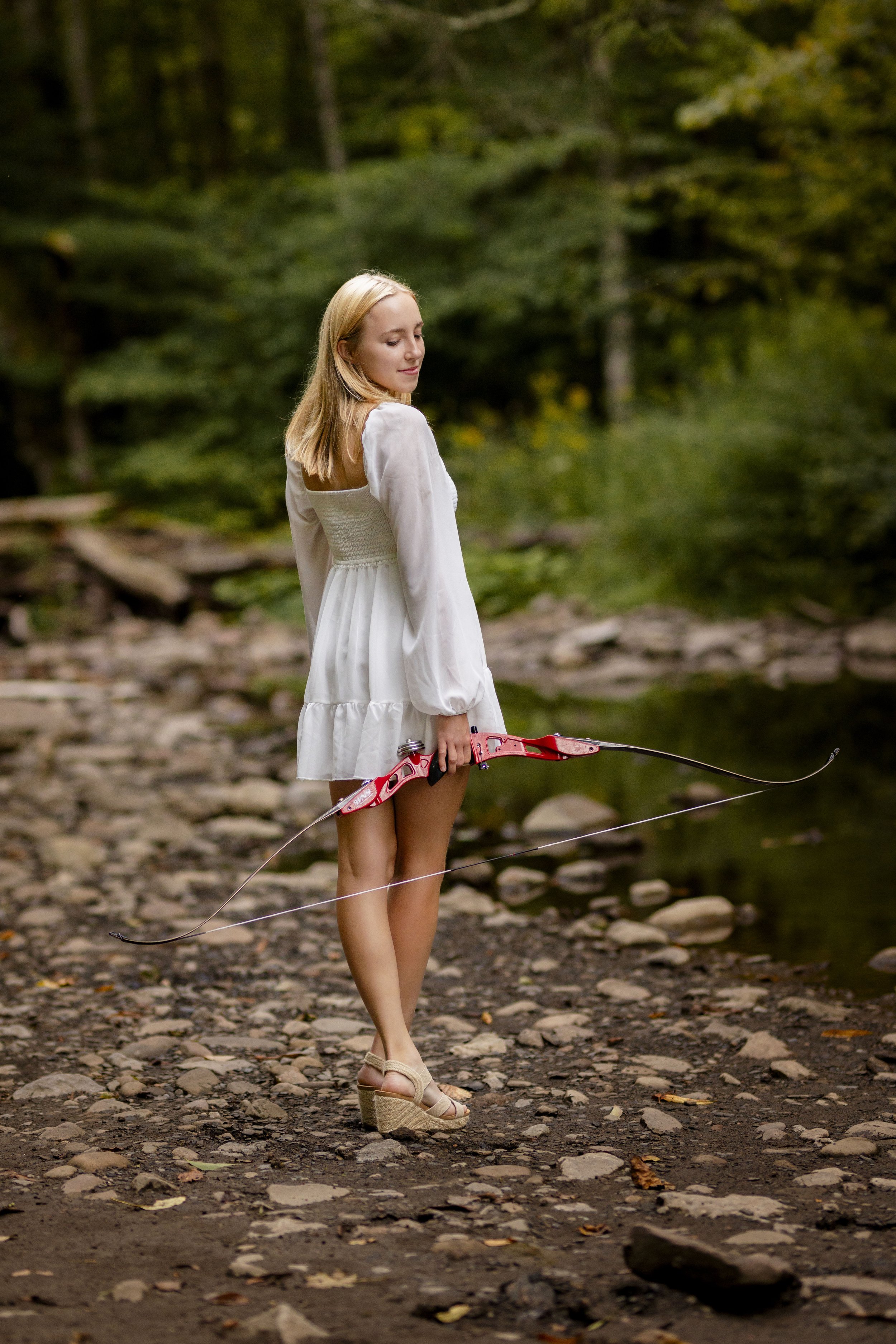 A young woman in a white dress and high-heeled sandals standing on a rocky forest path, holding a pink bow and arrow, with a calm expression and eyes closed.