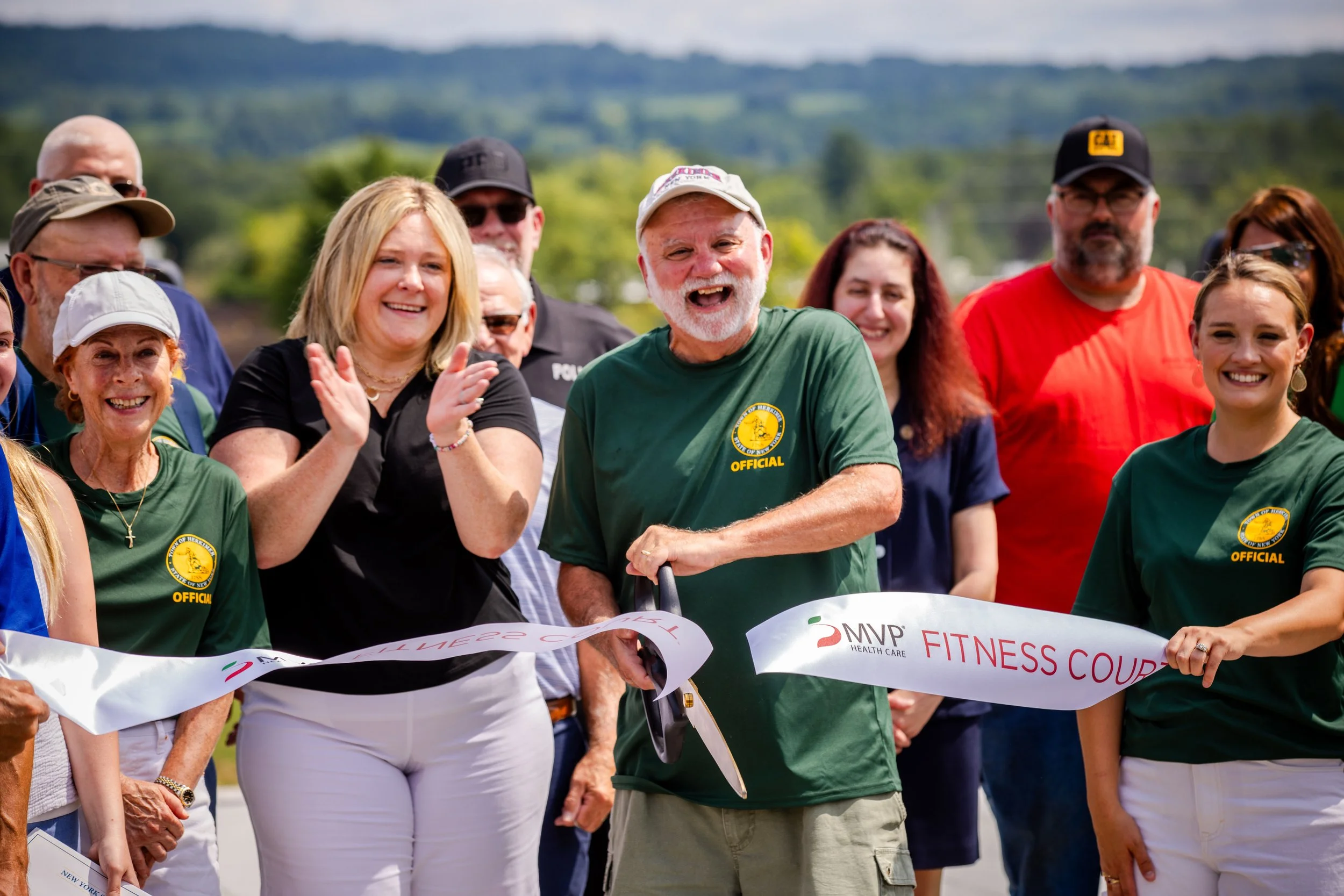 Person cutting a ribbon at a community event with people clapping and smiling, outdoors with green landscape in background.