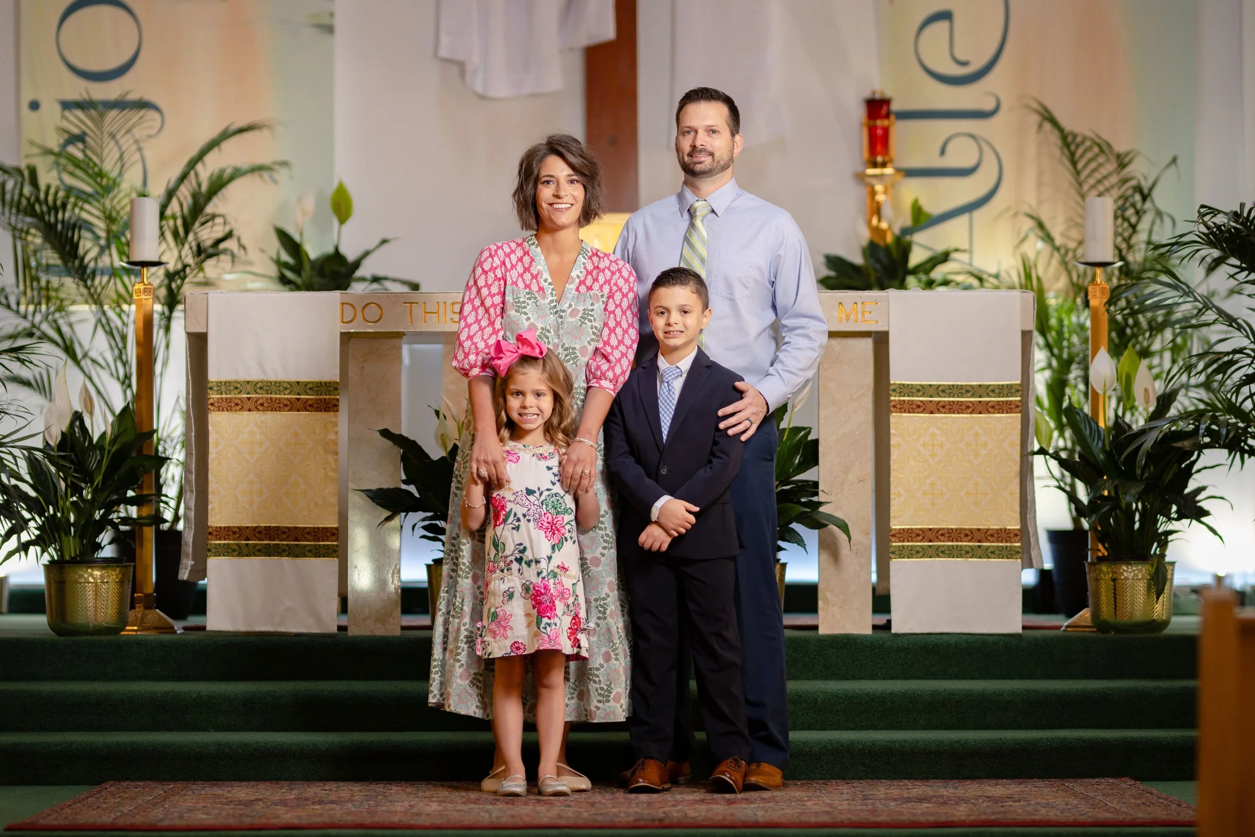 A family standing on the steps of a church altar during a religious ceremony. The woman is wearing a pink floral dress, the man is in a blue shirt and tie, the girl is in a floral dress with a pink bow, and the boy is in a suit and tie. The altar is decorated with plants, candles, and religious symbols.