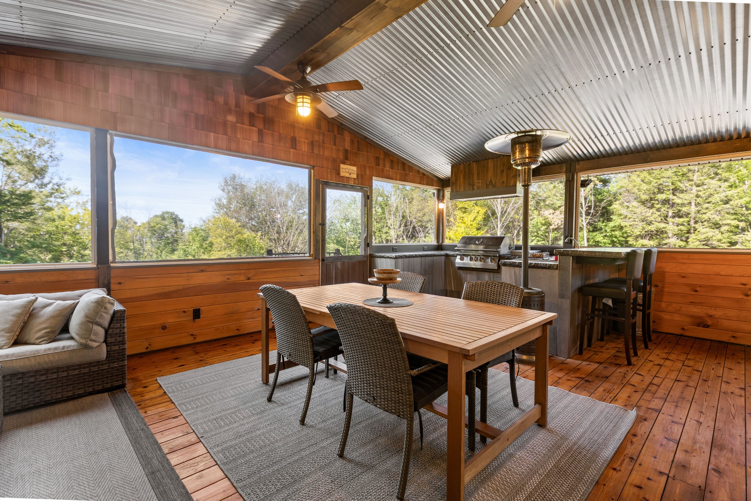Wooden screened porch with dining table, wicker chairs, a ceiling fan, a patio heater, and a grill, overlooking lush green trees.