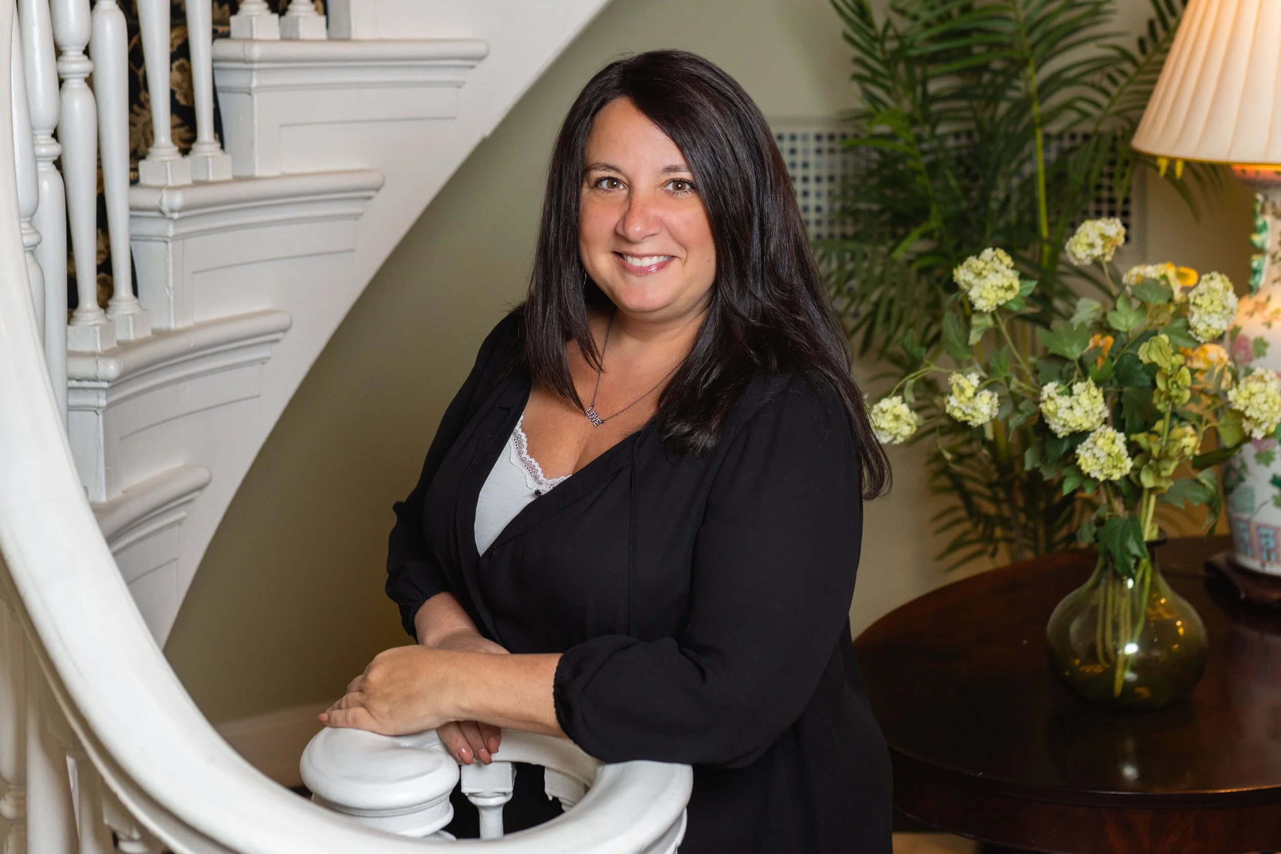 A woman with dark brown hair in a black top standing beside a white staircase, smiling, with a wooden table and green plant with white flowers in the background.