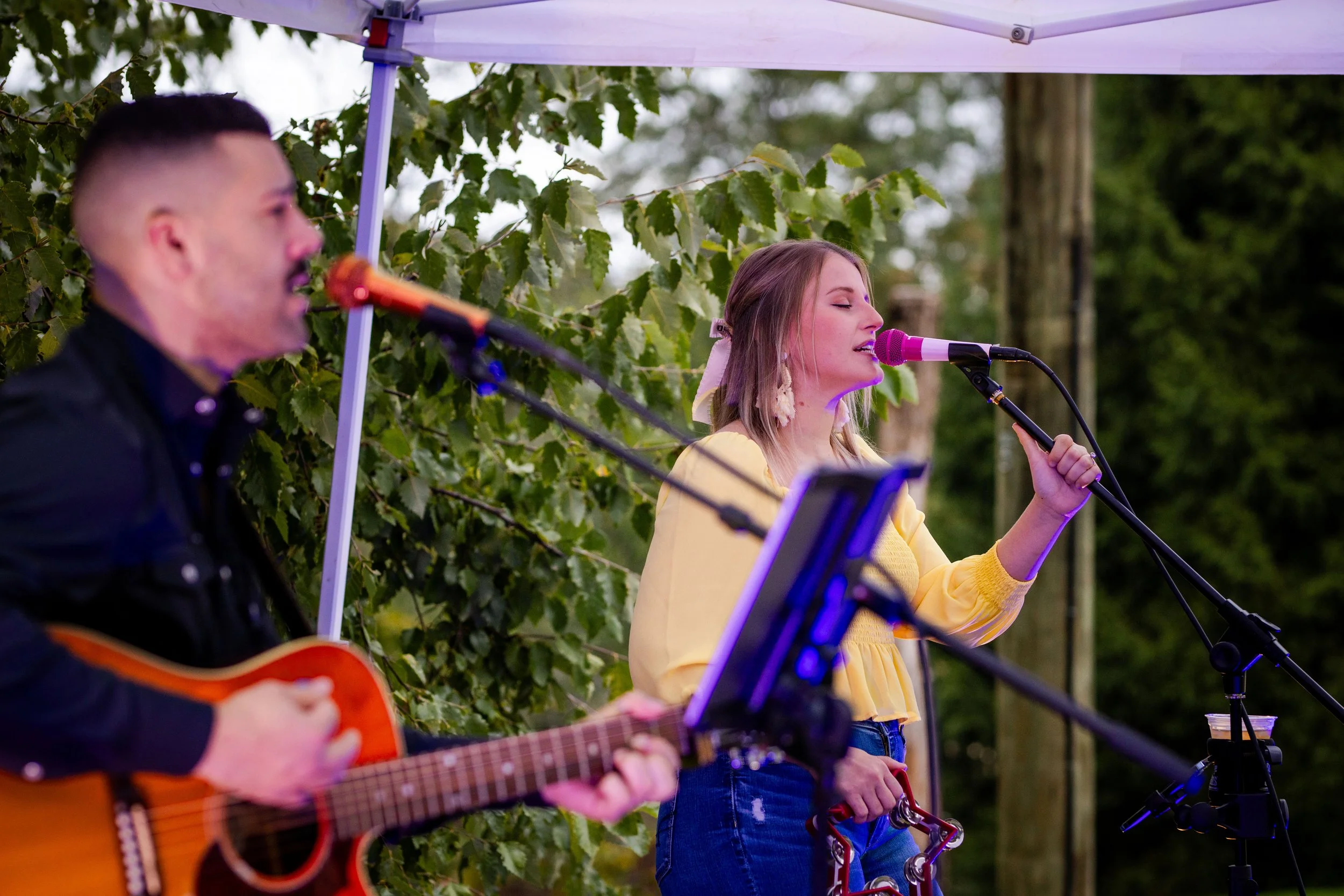 A woman in a yellow top singing into a microphone while holding a tambourine, accompanied by a man playing an acoustic guitar, performing outdoors under a white canopy with green foliage in the background.