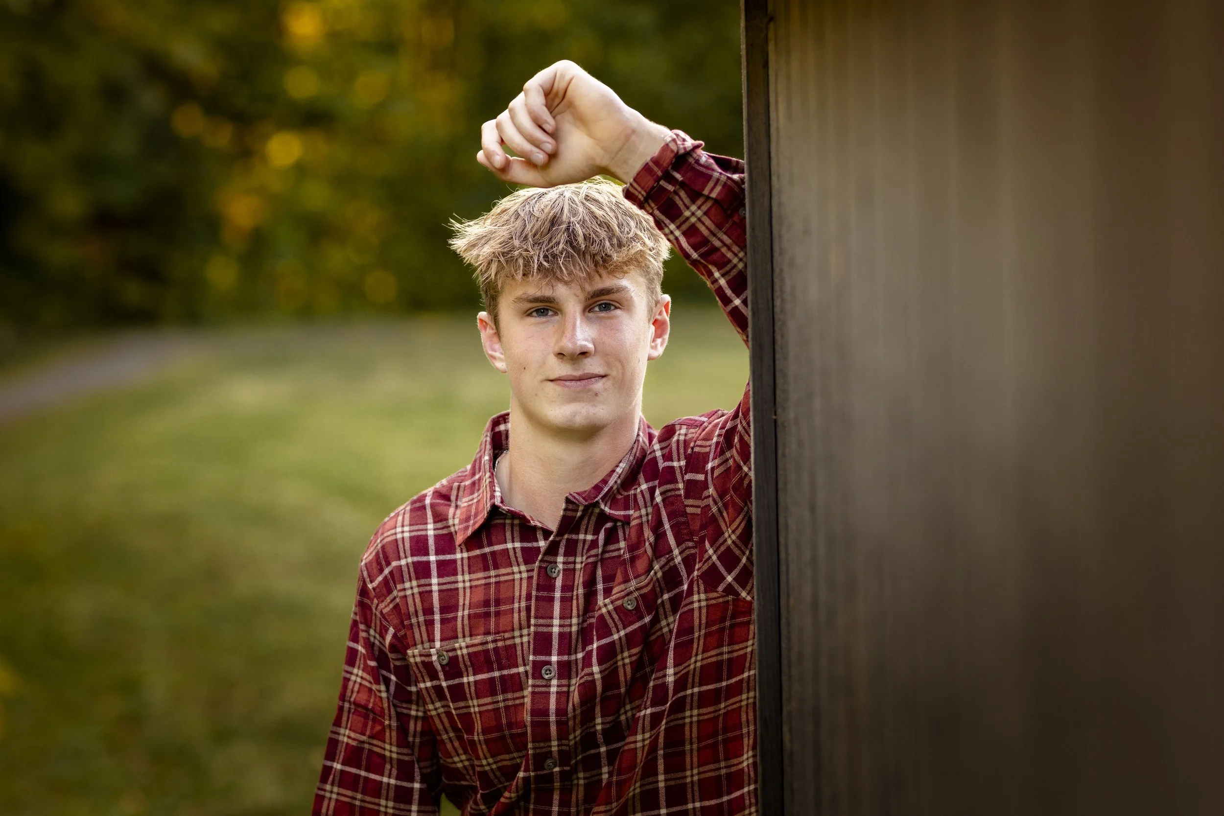 Young man with light brown hair wearing a red plaid shirt standing outdoors near a wooden structure, with a blurred background of trees and grass.
