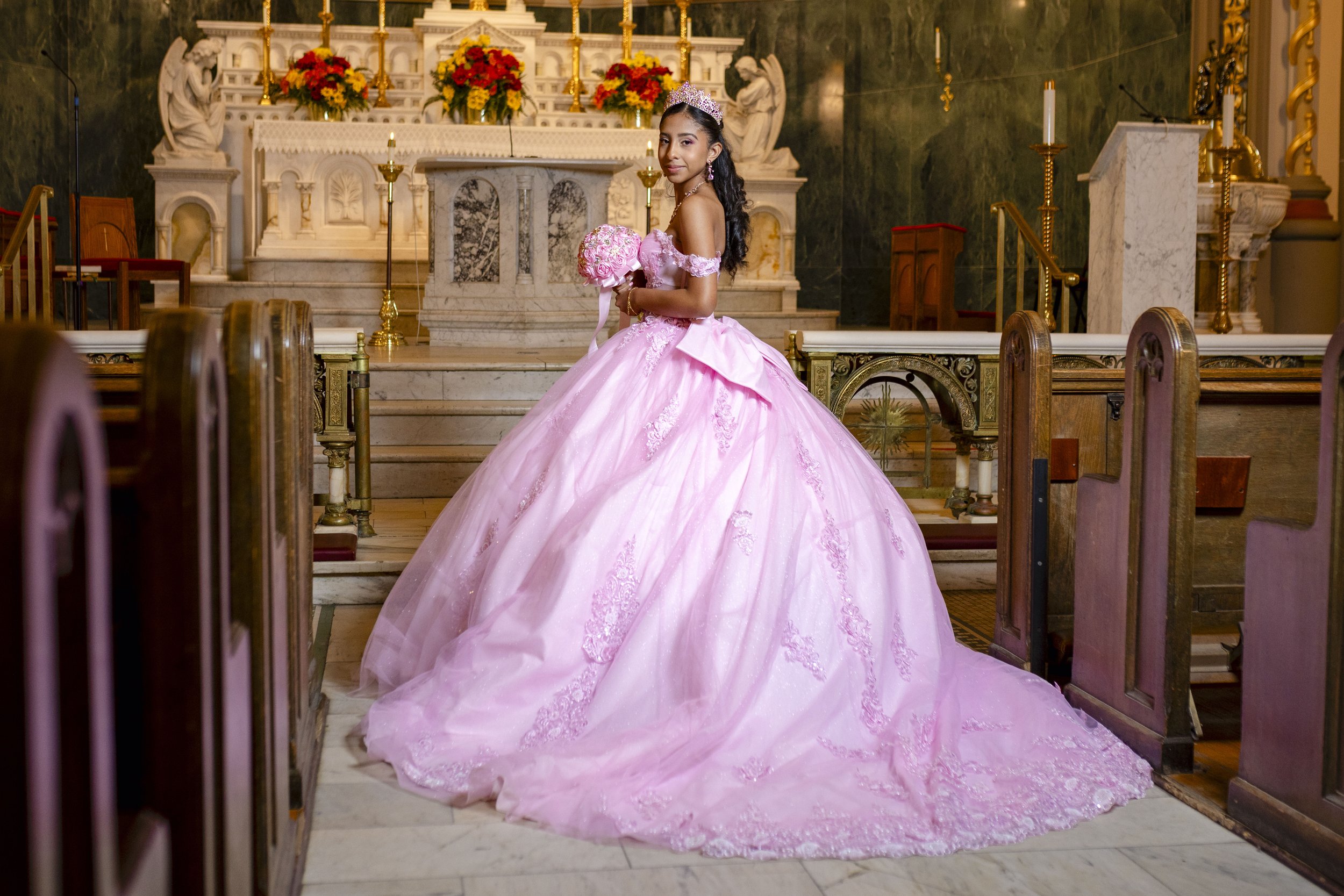 A young girl in a pink ball gown with lace and floral embroidery, wearing a tiara, stands inside a church holding a bouquet of flowers, with an ornate altar decorated with flowers behind her.