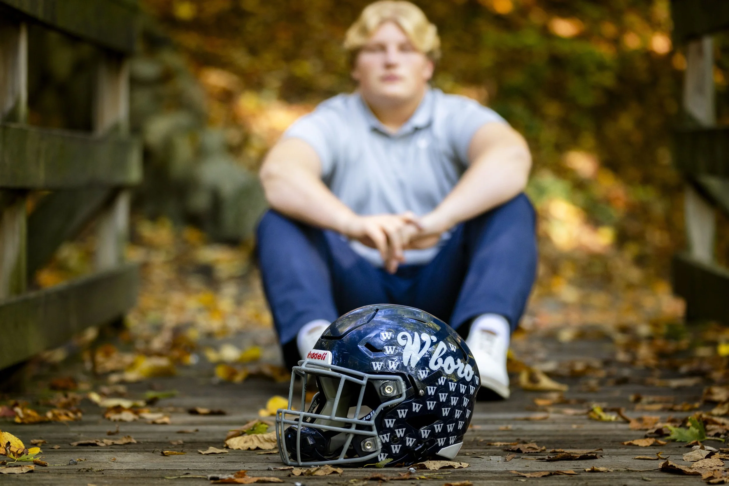 A young man sitting on a wooden bridge surrounded by autumn leaves, with a Seahawks football helmet placed in front of him.