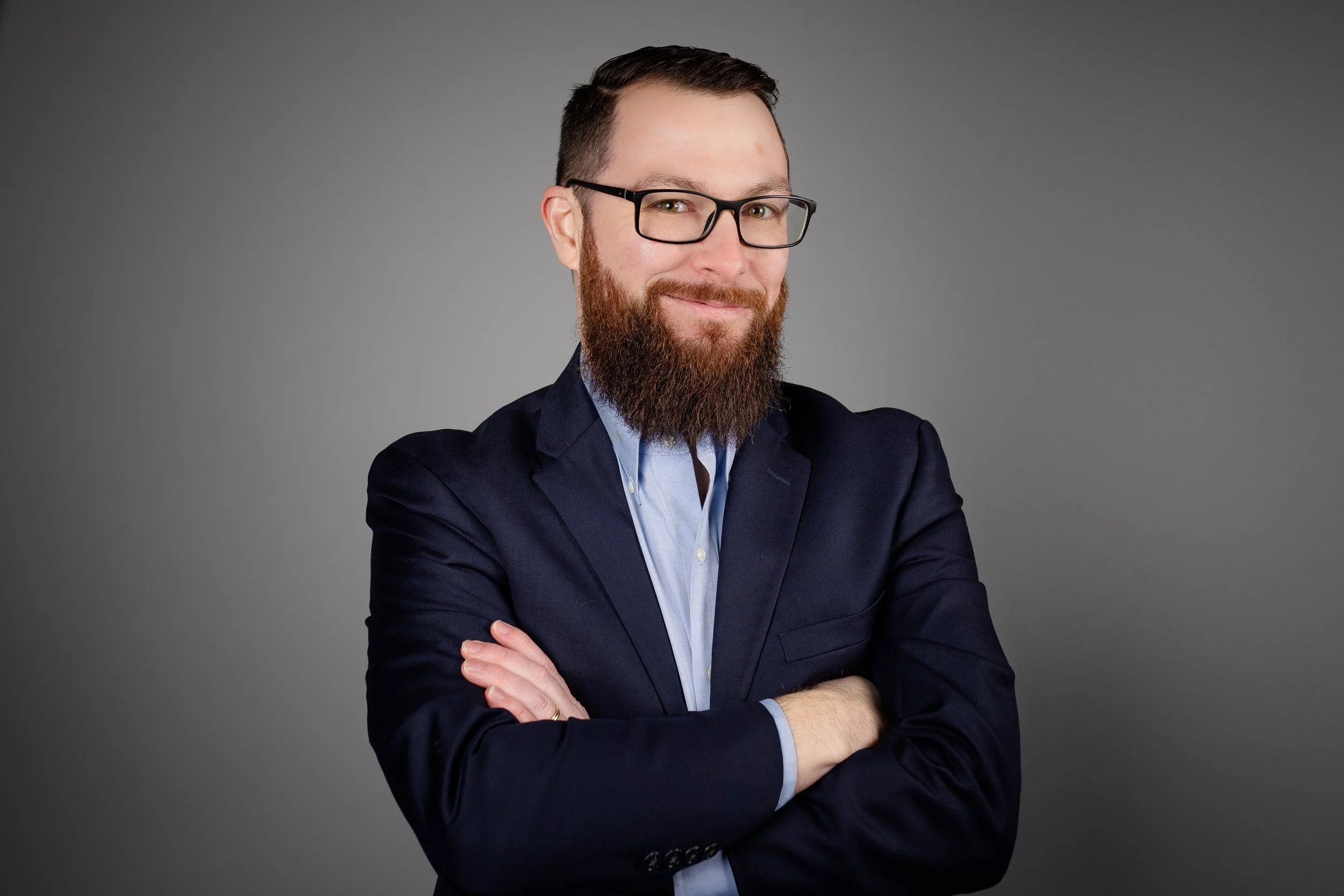 A man with a beard and glasses wearing a navy blazer and light blue shirt, smiling with arms crossed, standing against a gray background.
