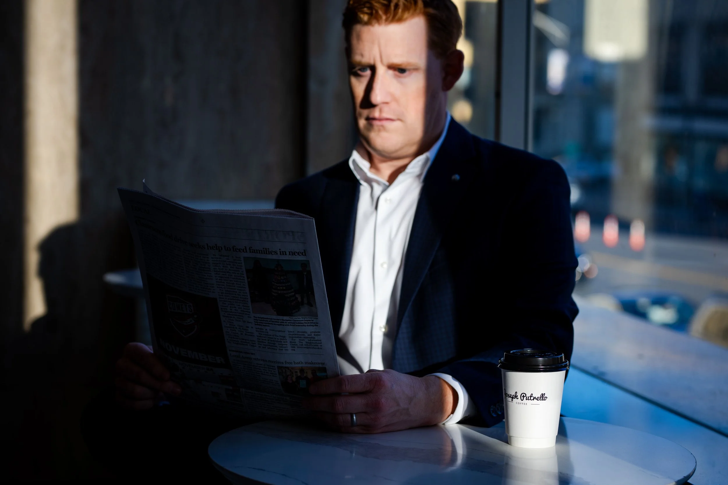 A man in a dark suit and white shirt sitting at a table by a window, reading a newspaper, with a coffee cup on the table.