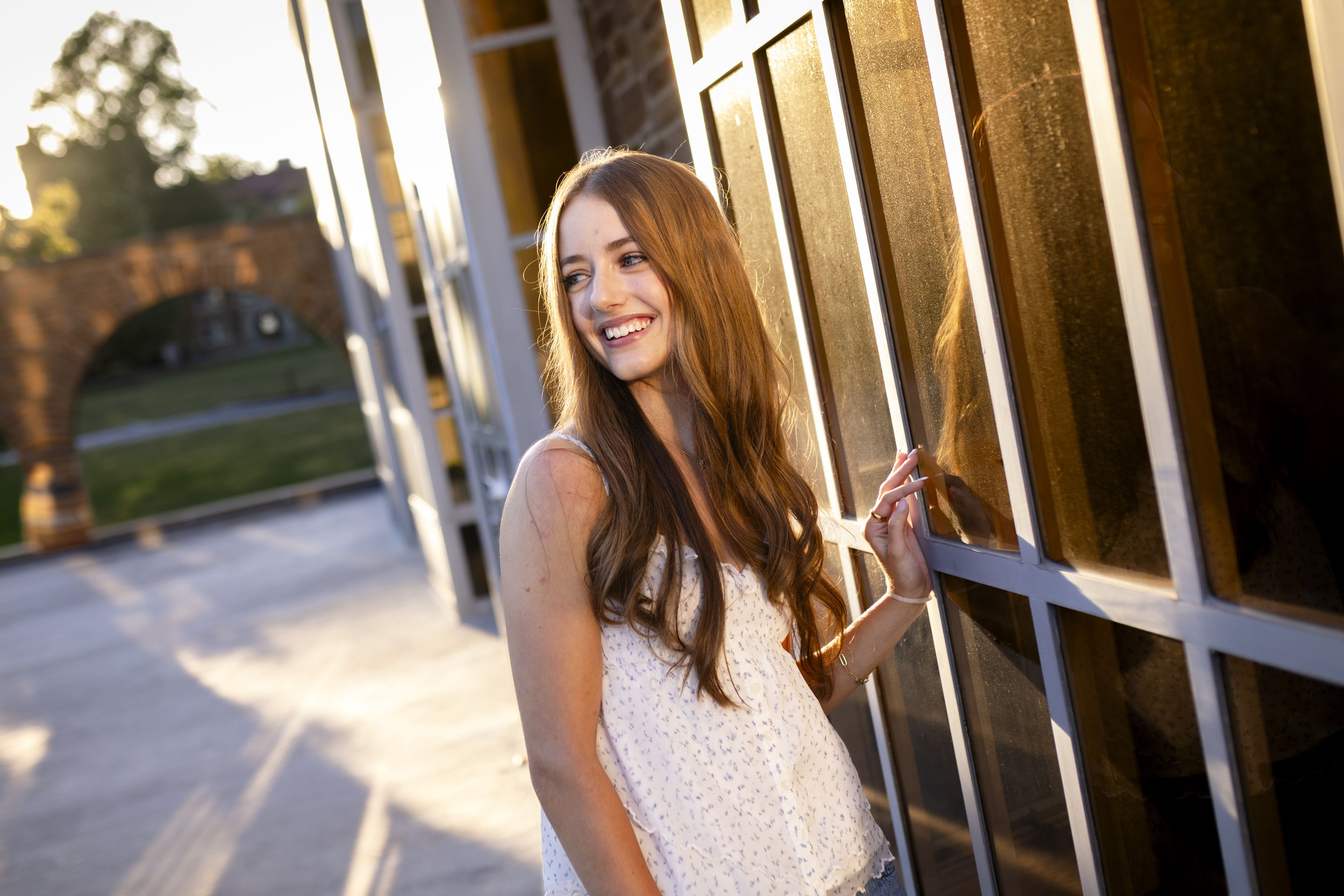 A young woman with long red hair smiling while standing outdoors at sunset next to a building with large windows.