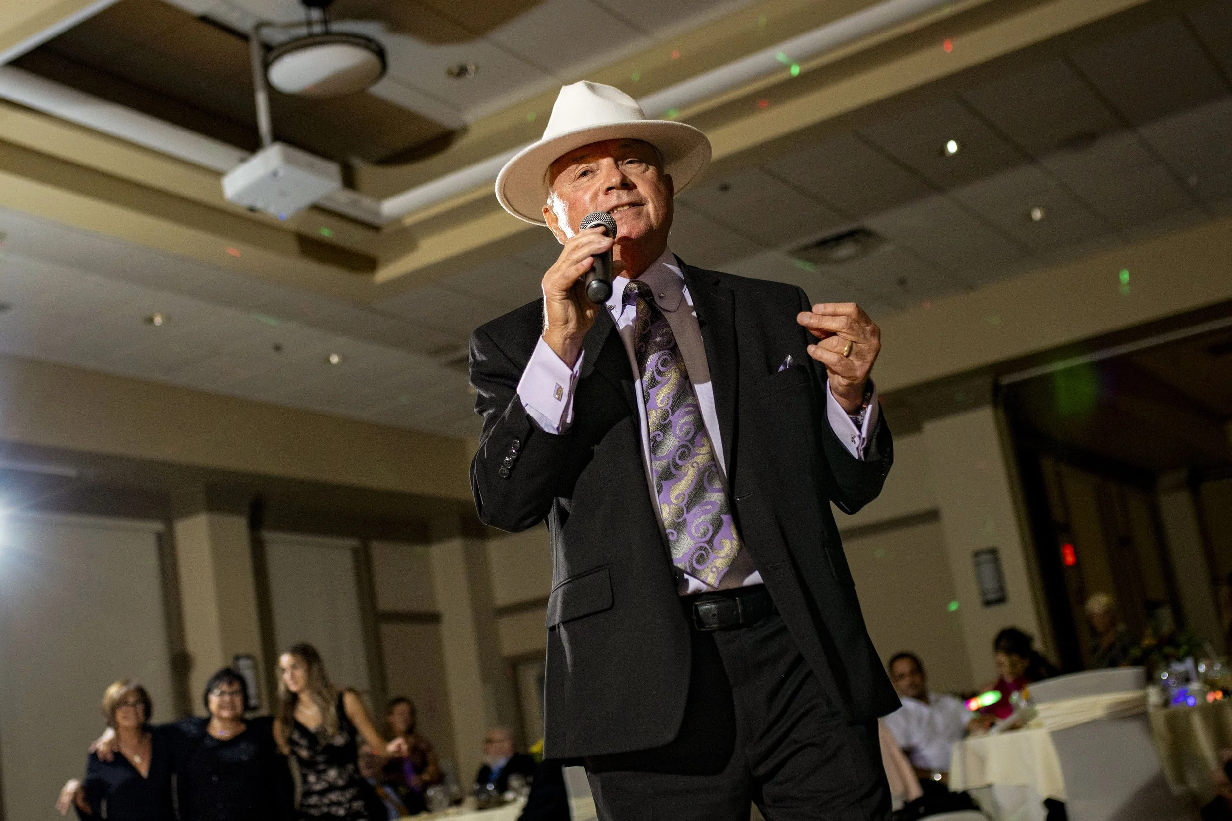 An elderly man in a black suit, dress shirt, and patterned tie, wearing a white wide-brimmed hat, holding a microphone and speaking in a banquet hall with people seated at tables in the background.