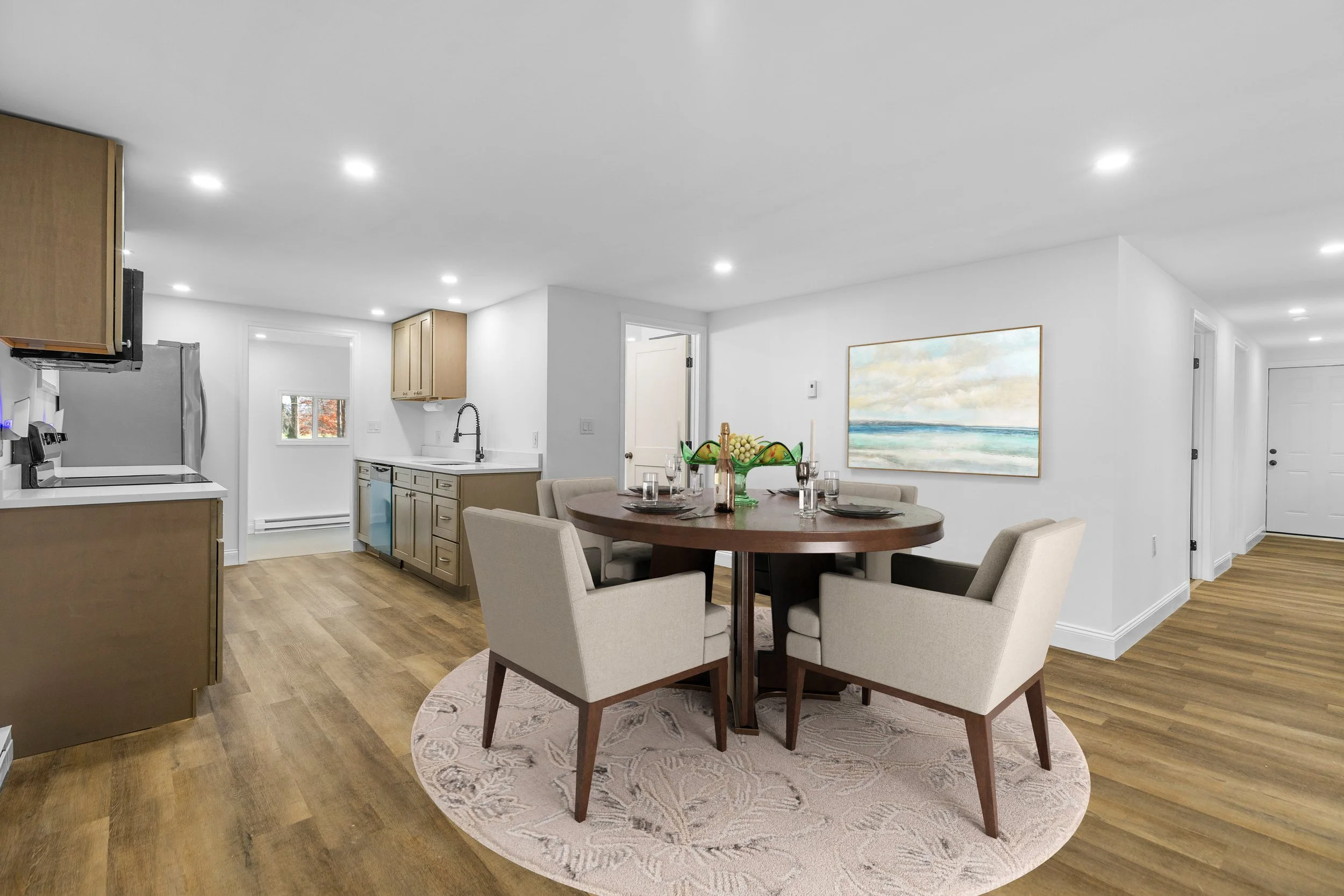 A dining area with a round wooden table set for four people, with beige upholstered chairs, and a fruit bowl centerpiece. The wall behind has a landscape painting, and to the left, there is a kitchen with wooden cabinets, a stainless steel refrigerator, and a small window.