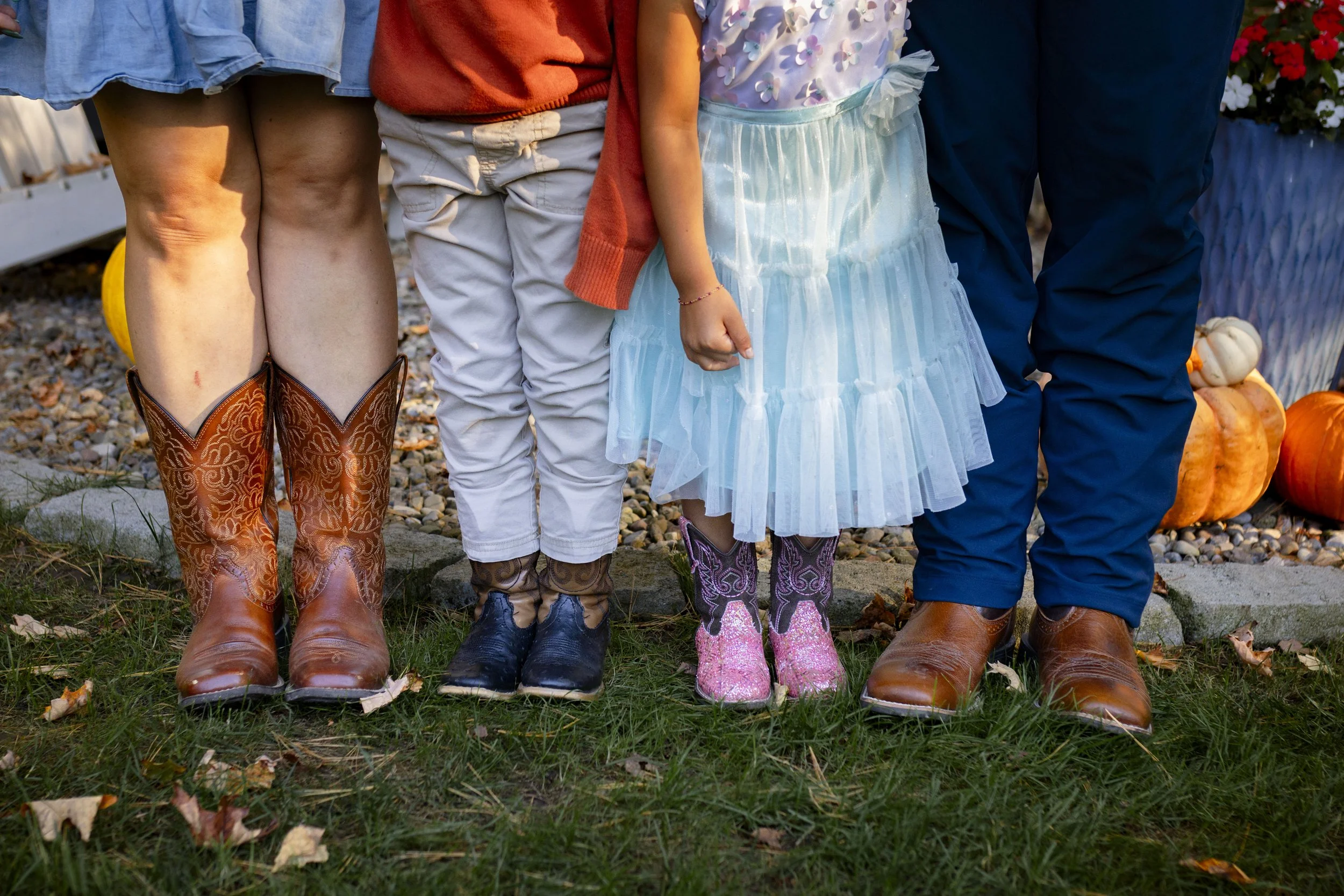 Close-up of four people standing outdoors, showing their lower legs and feet, all wearing cowboy boots. The scene includes a grassy area with pumpkins and flowers in the background.