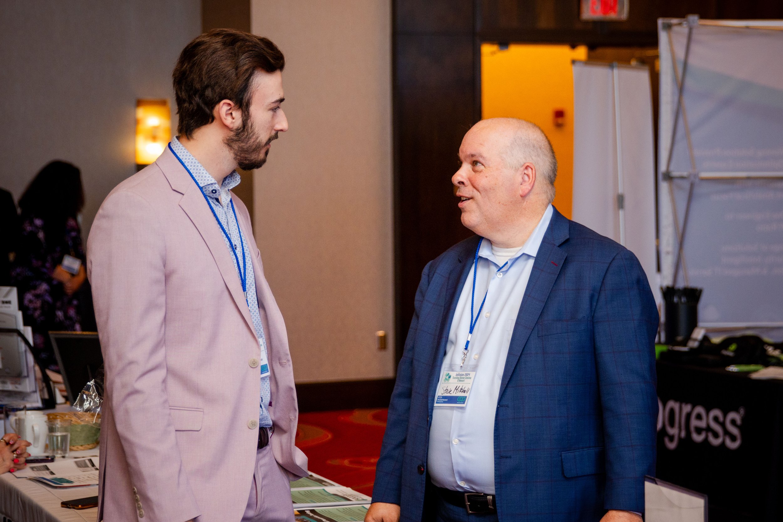 Two men having conversation at a conference, wearing business attire, with conference badges, in a room with display booths and a table with information pamphlets.