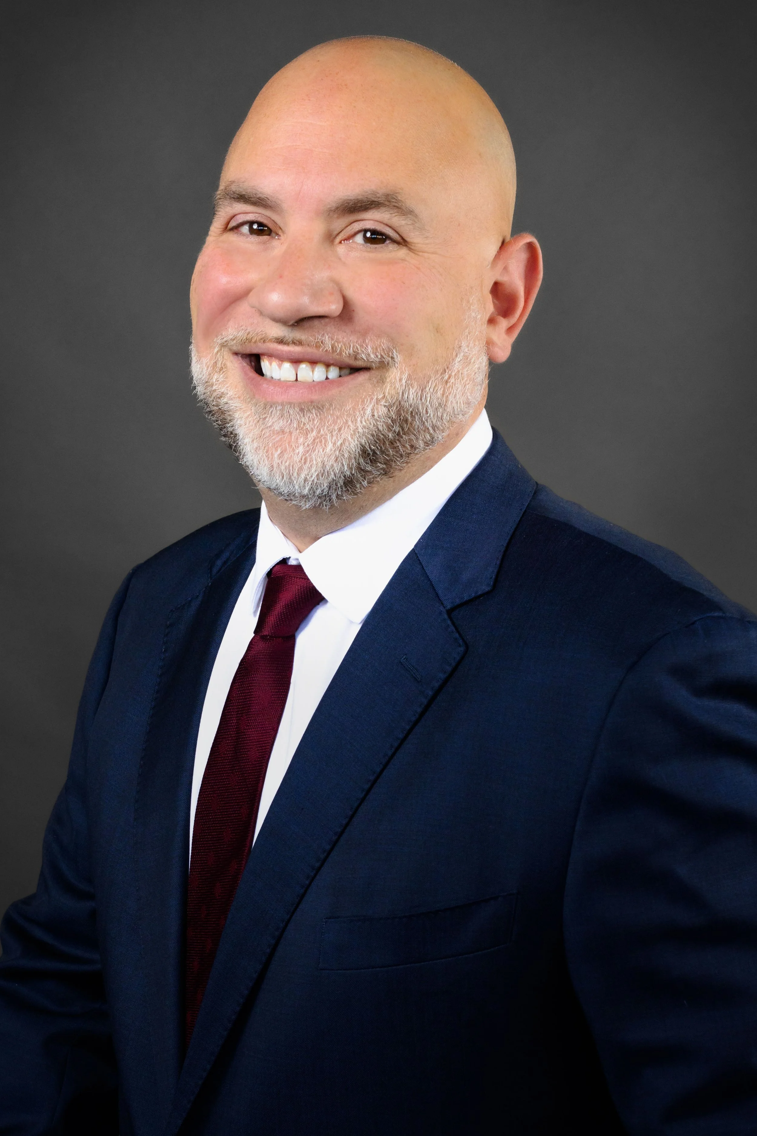 A smiling middle-aged man with a beard and bald head dressed in a dark suit, white shirt, and maroon tie, posed against a dark gray background.