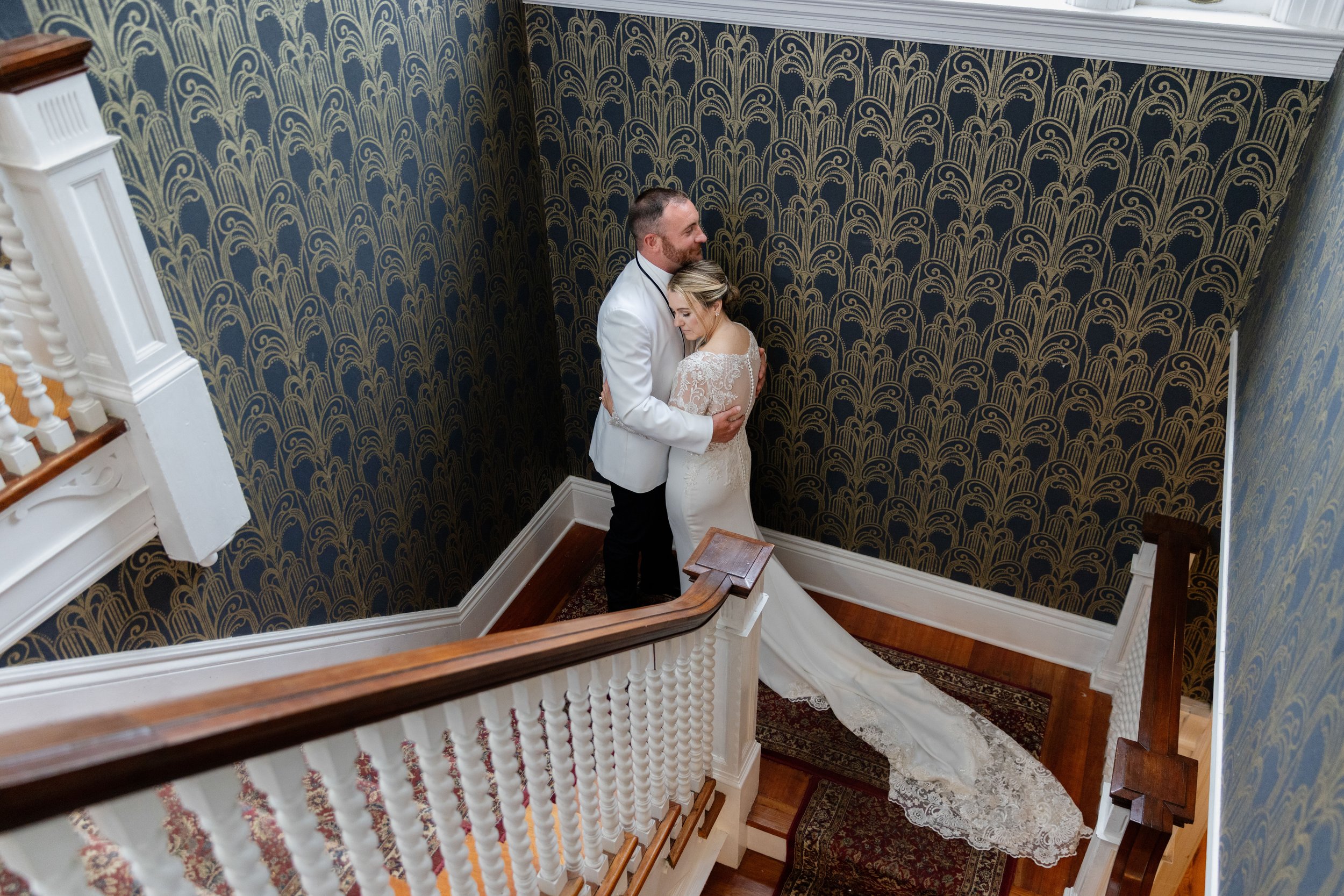 A bride and groom hug on a staircase, with ornate dark blue and gold wallpaper surrounding them, and hardwood flooring under a patterned rug.