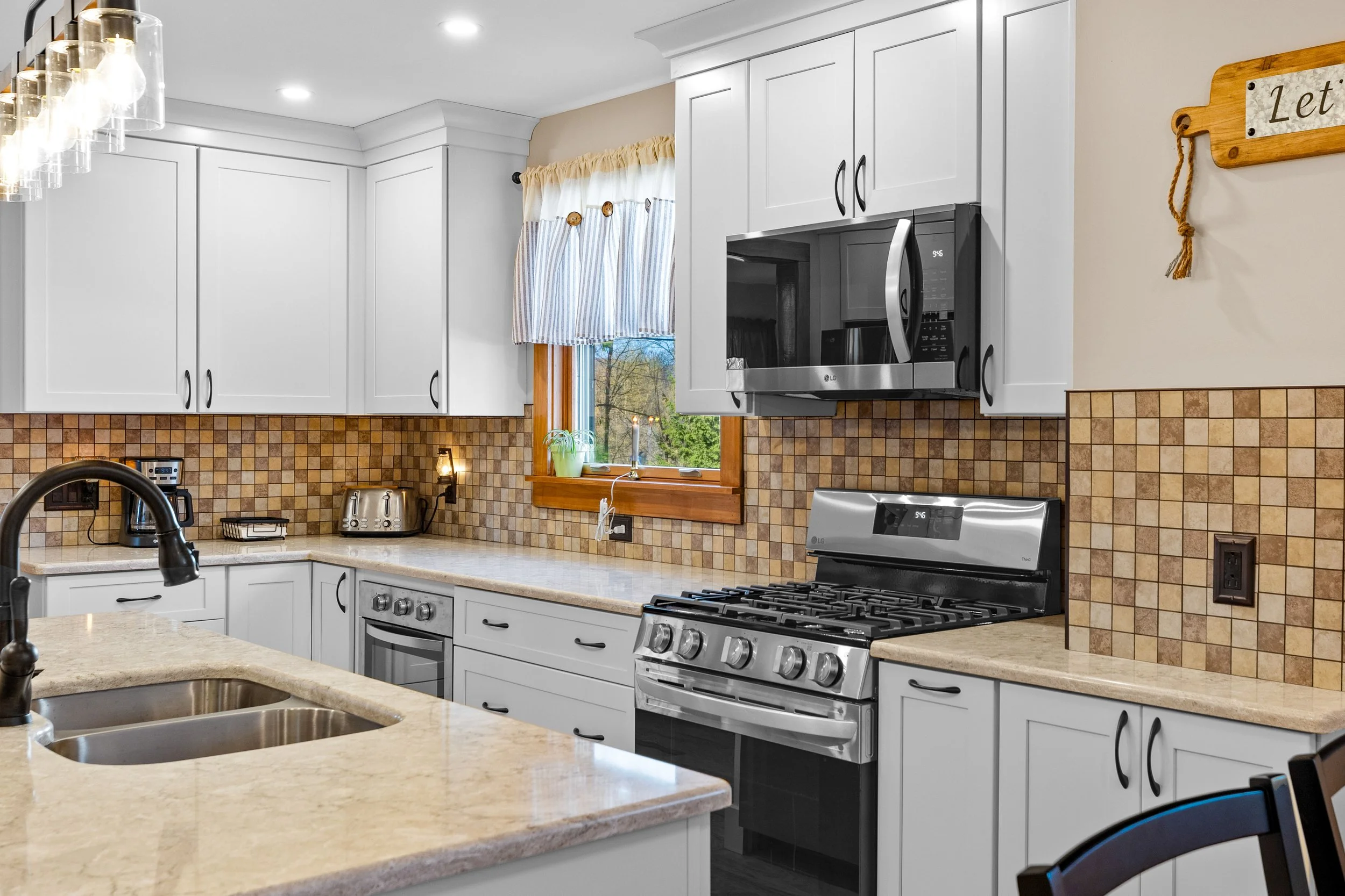 Kitchen with white cabinets, beige countertops, a stainless steel oven and microwave, a tiled backsplash, a window with a striped curtain, and various small appliances.