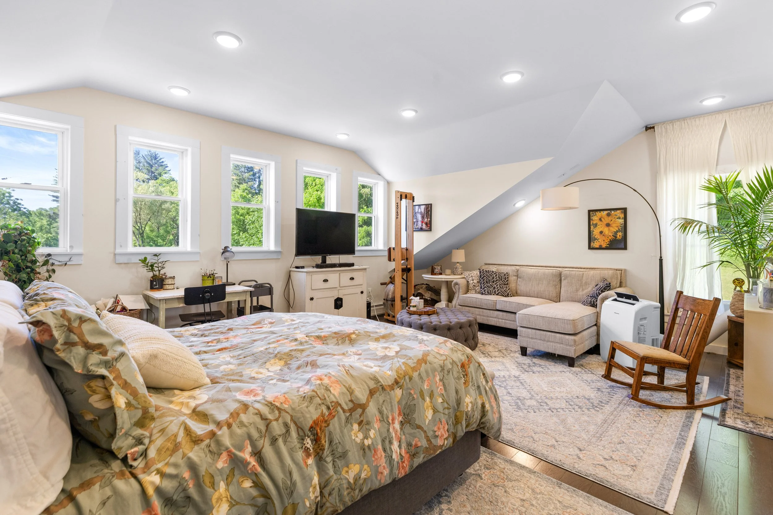 Bright bedroom with multiple large windows, a bed with floral bedding, a small desk with a chair, a television on a white dresser, and a sitting area with a sectional sofa, rocking chair, and large green plant.
