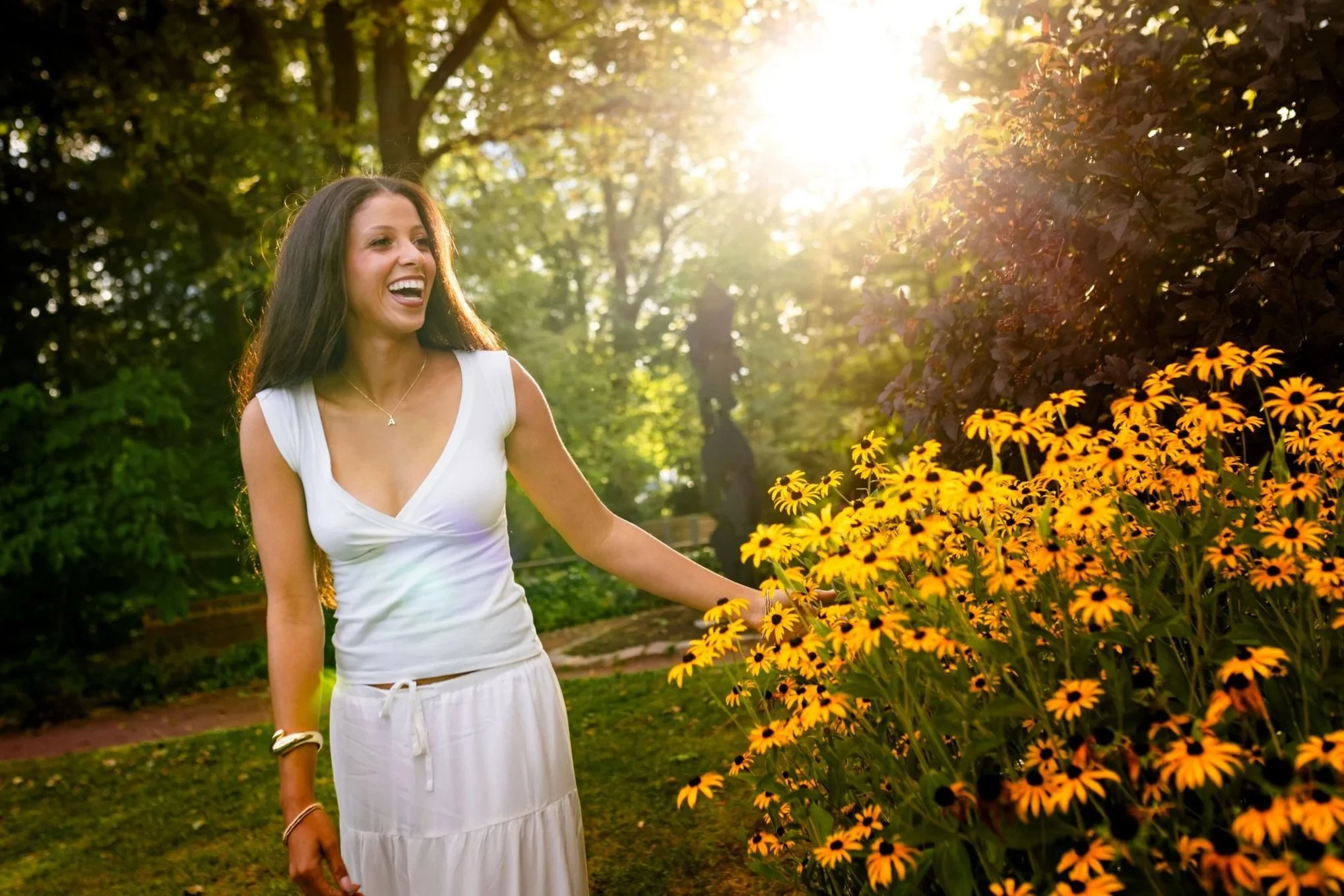 A young woman in a white dress standing outdoors near a bush of yellow flowers, smiling and enjoying the sunlight.