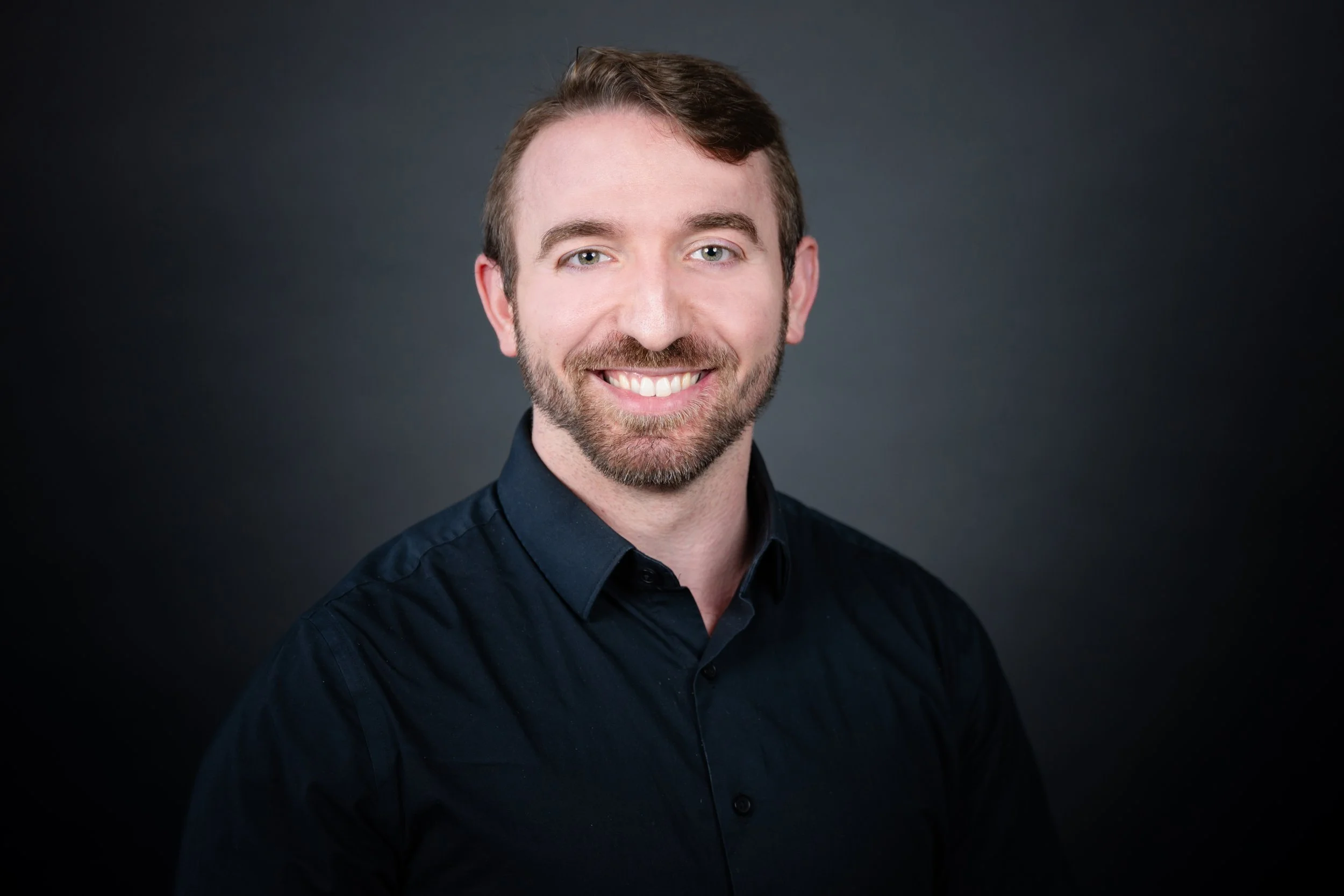 Portrait of a smiling man with light brown hair, beard, wearing a black shirt, against a dark gradient background.