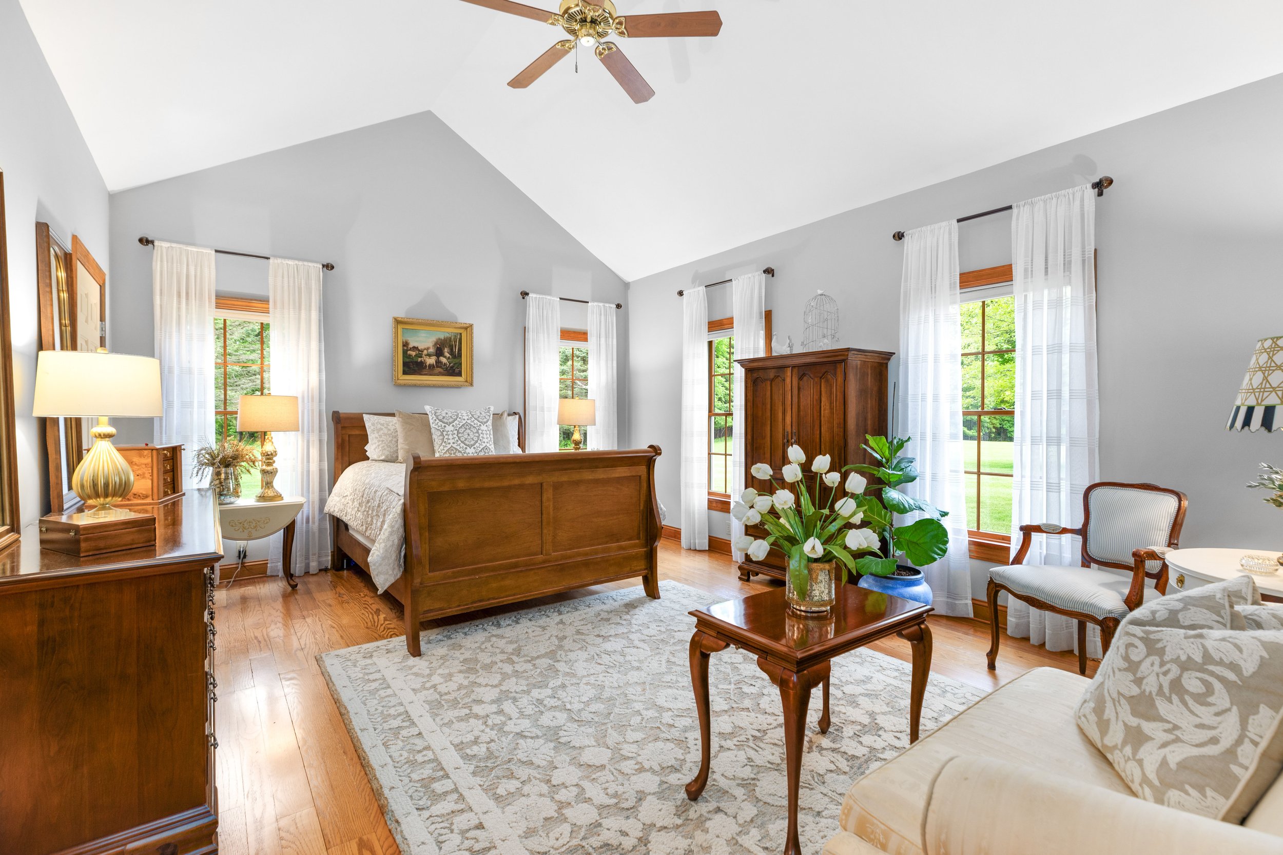 Bright bedroom with wooden furniture, white curtains, a ceiling fan, and a floral rug.