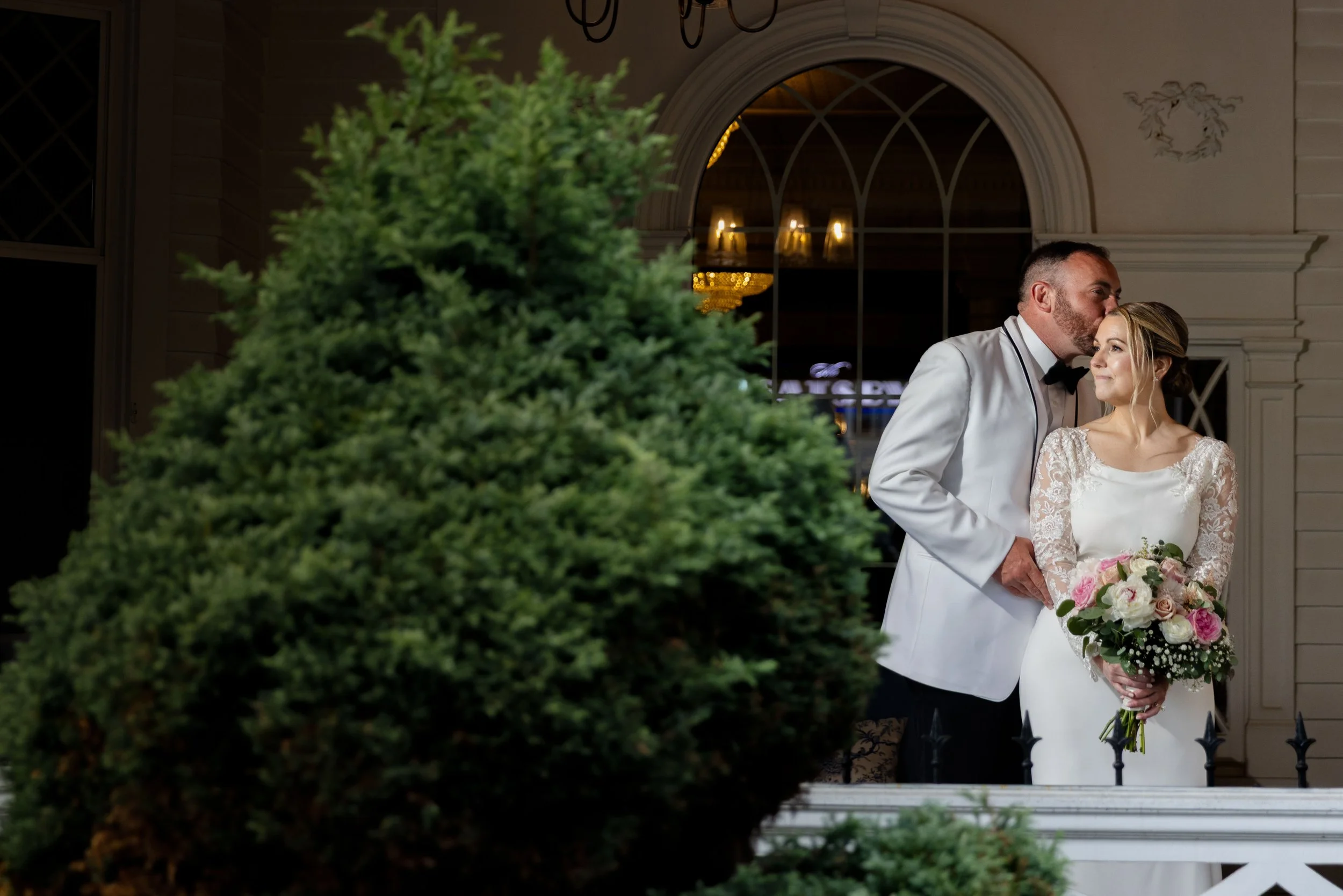 A bride and groom stand together on a balcony, with the groom kissing the bride's forehead. The bride is holding a bouquet of roses and white flowers, and she is wearing a white wedding dress with lace sleeves. The groom is dressed in a white tuxedo 