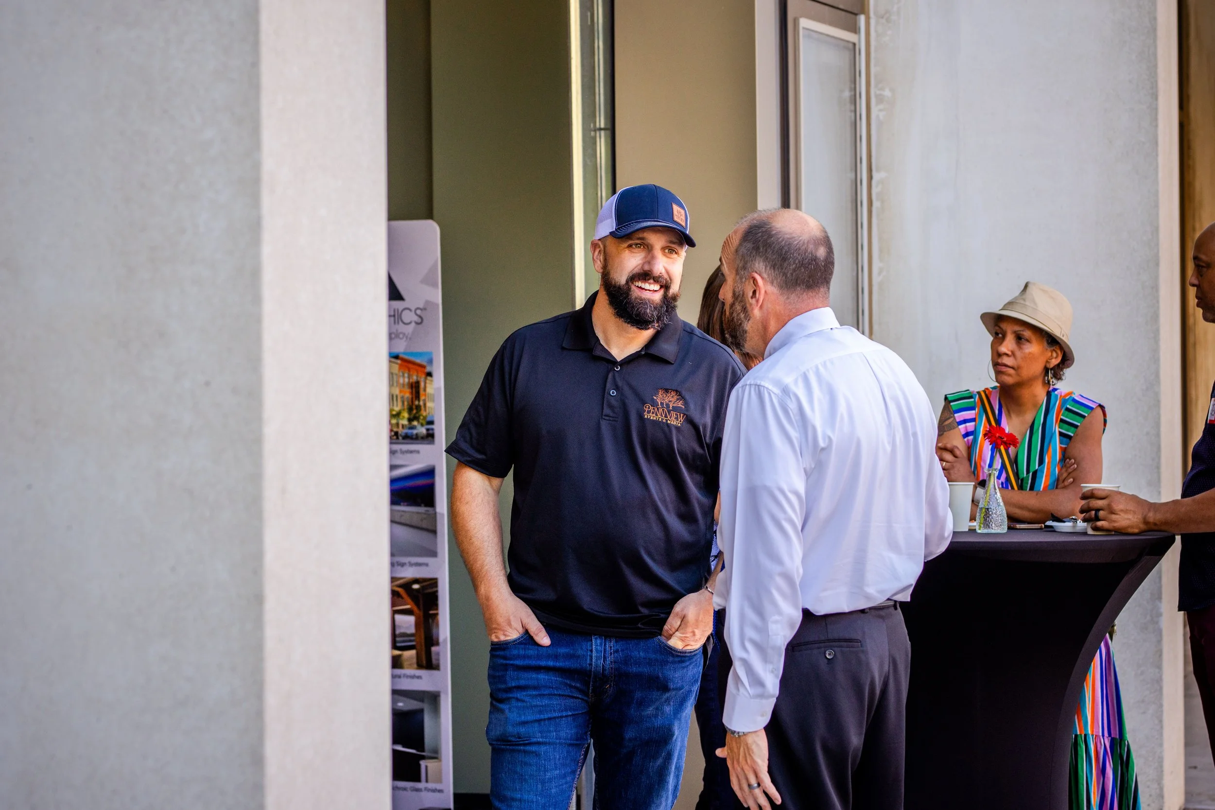 Group of people socializing at an outdoor event, with two men in conversation, one wearing a black polo shirt and baseball cap, and the other in a white dress shirt. In the background, a woman with a striped dress and beige hat stands by a table.