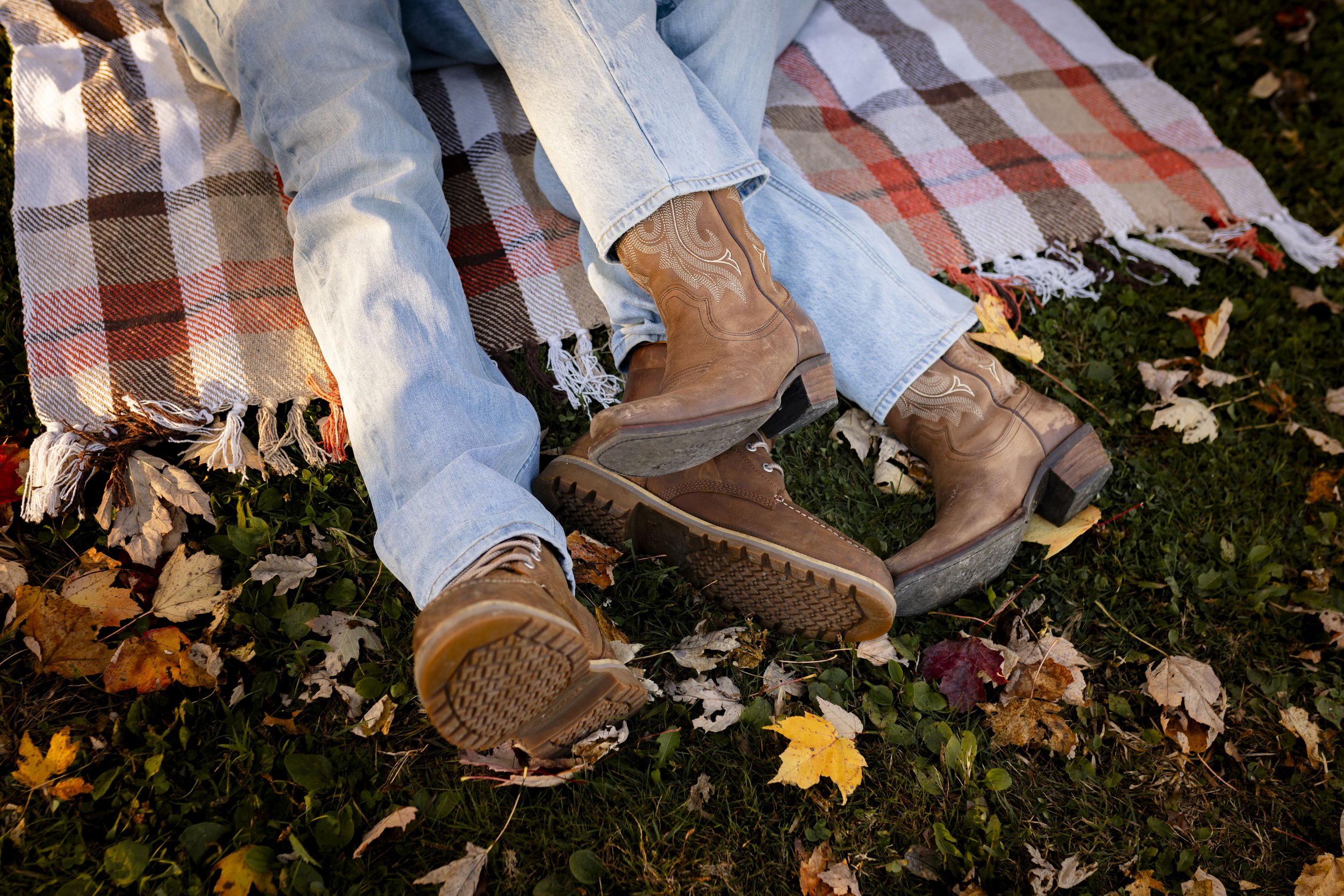 Two people are sitting on a plaid blanket on the grass with fallen autumn leaves, wearing jeans and cowboy boots.