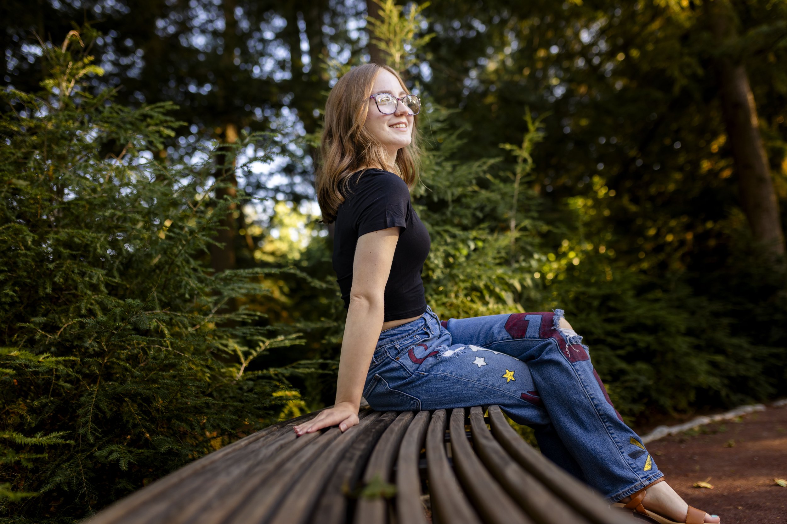 A young woman with glasses, long brown hair, wearing a black t-shirt and distressed denim jeans, sitting on a wooden park bench surrounded by green trees in sunlight.