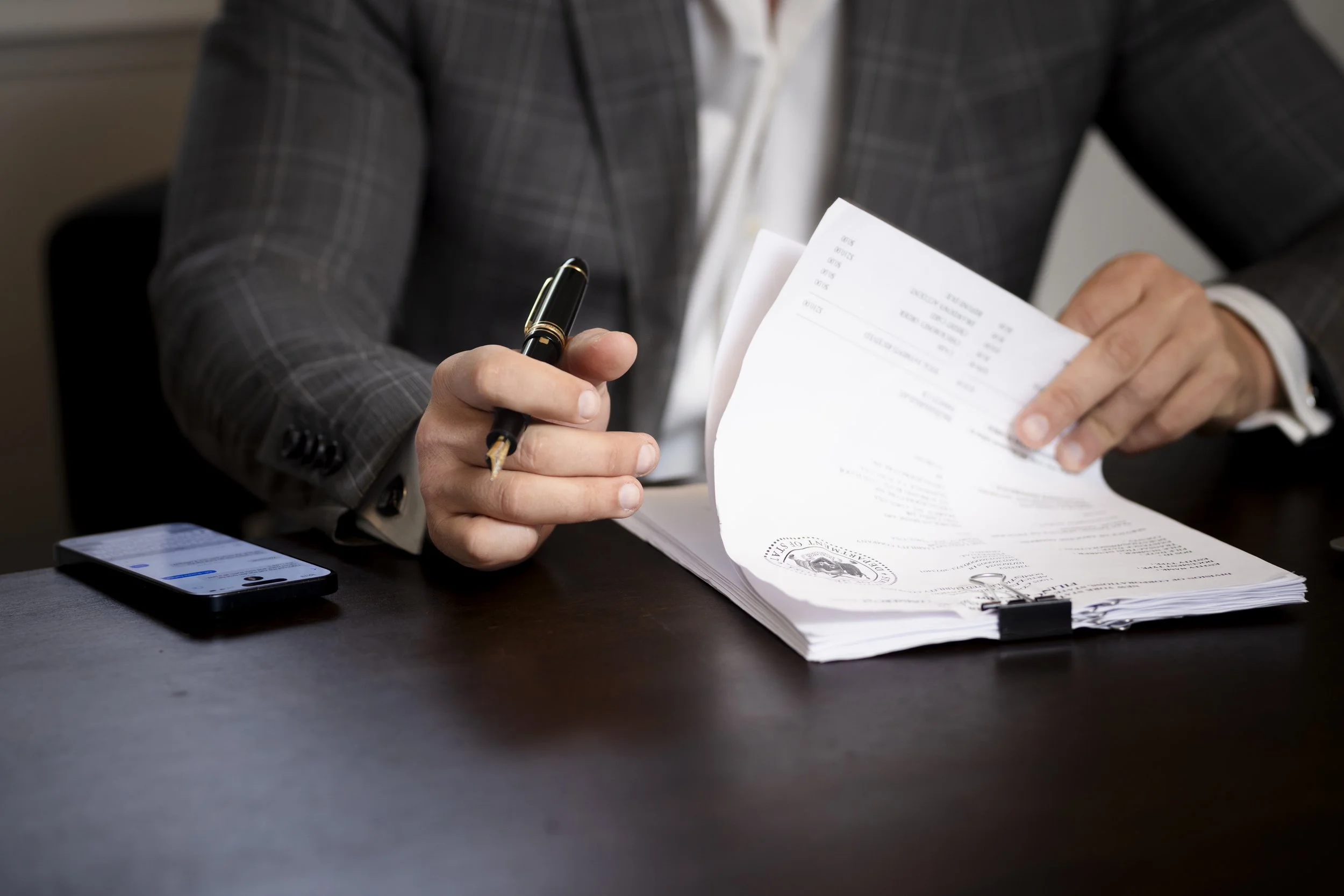 Close-up of a person in a grey plaid suit holding a pen and reviewing a stack of documents on a dark surface, with a smartphone nearby.