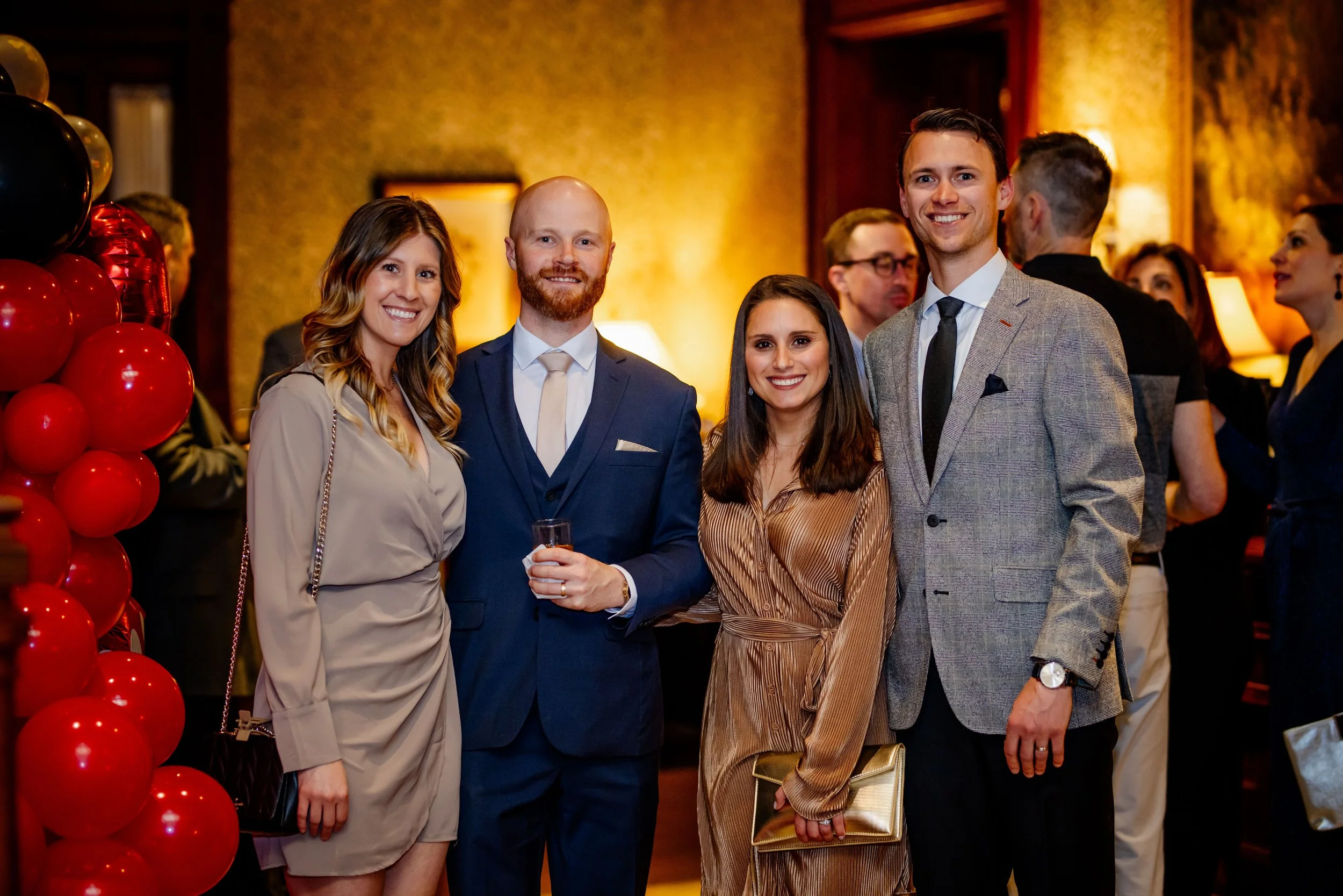 Group of four people dressed in formal attire at a celebration event, standing indoors near red and black balloon decorations.