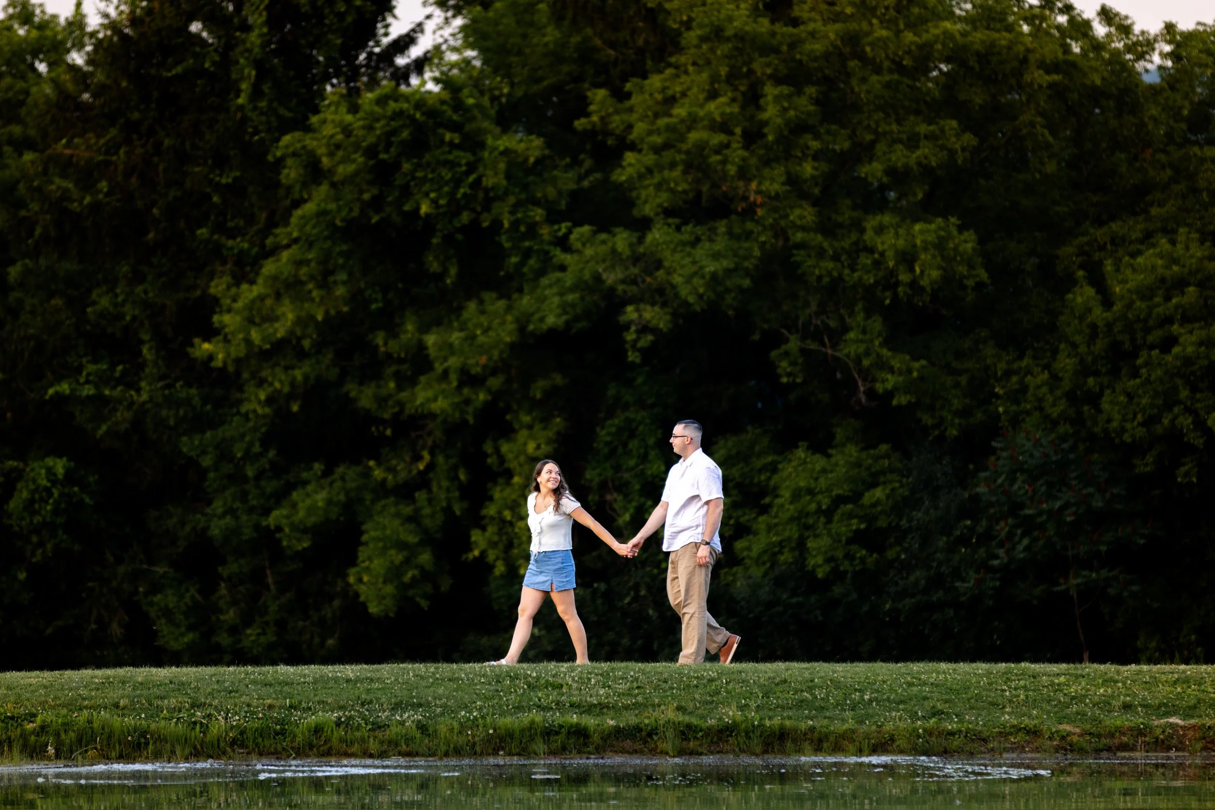 A couple holding hands and smiling while walking near a body of water with a background of dense green trees.