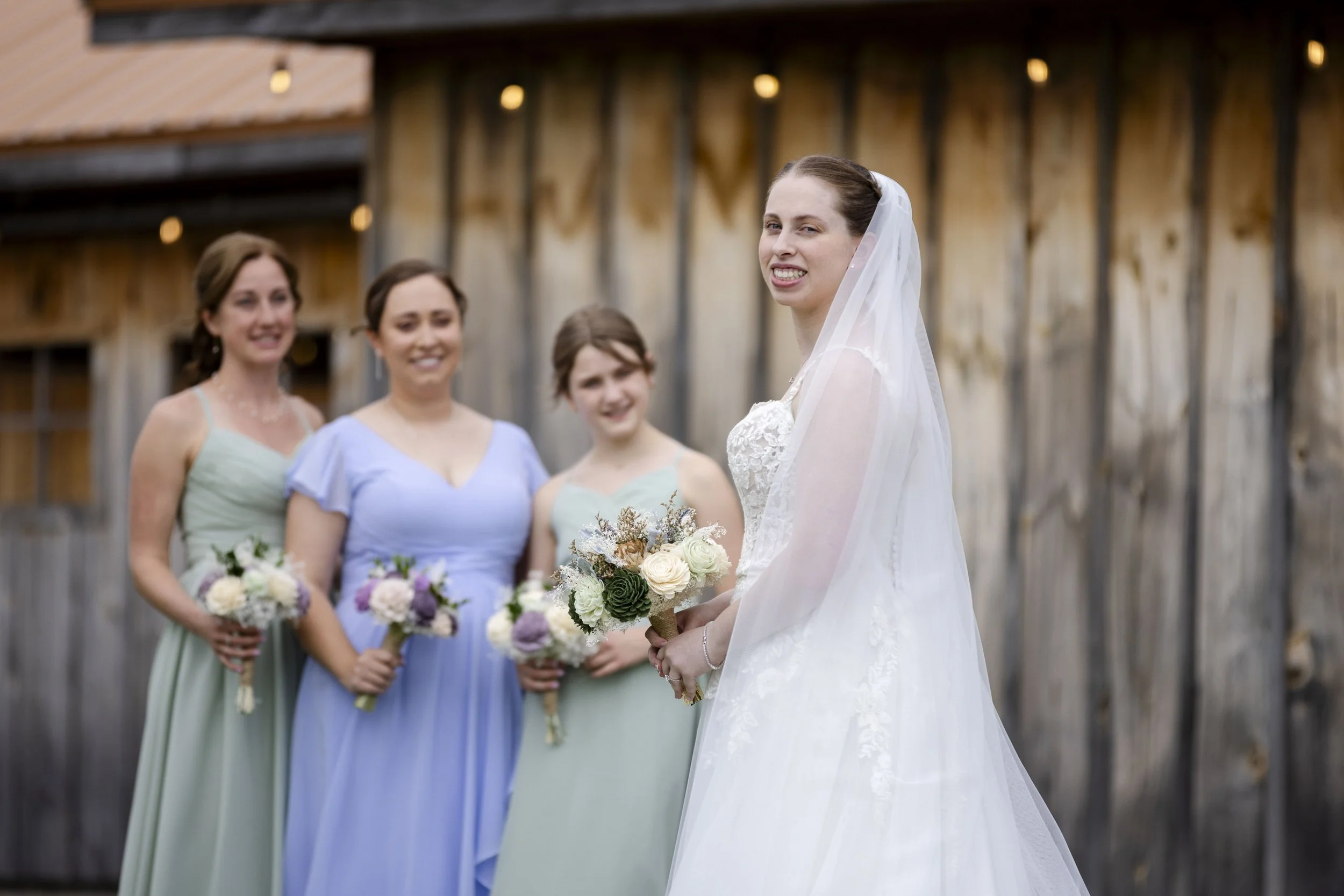 A bride holding a bouquet of flowers standing outside with three bridesmaids, all smiling, with a rustic wooden barn background.