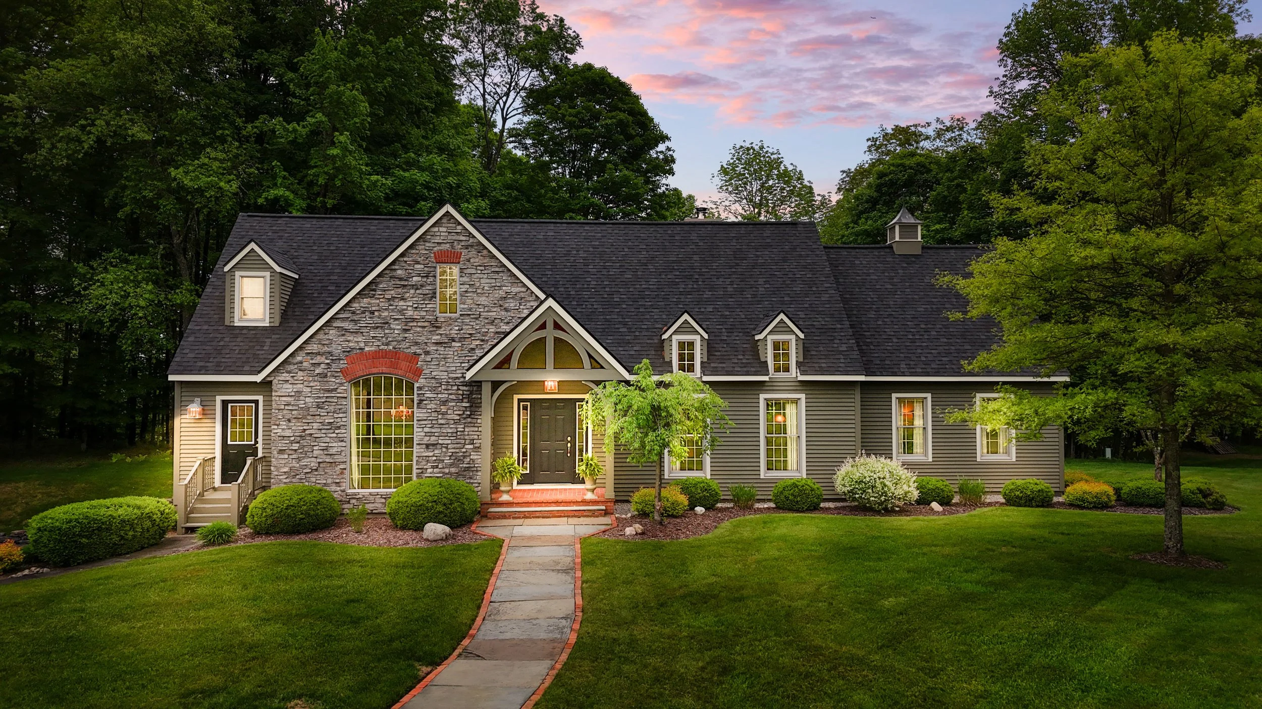 A large, elegant house with stone and siding exterior, multiple small gabled dormer windows, and a dark shingle roof, set in a landscaped yard at dusk.