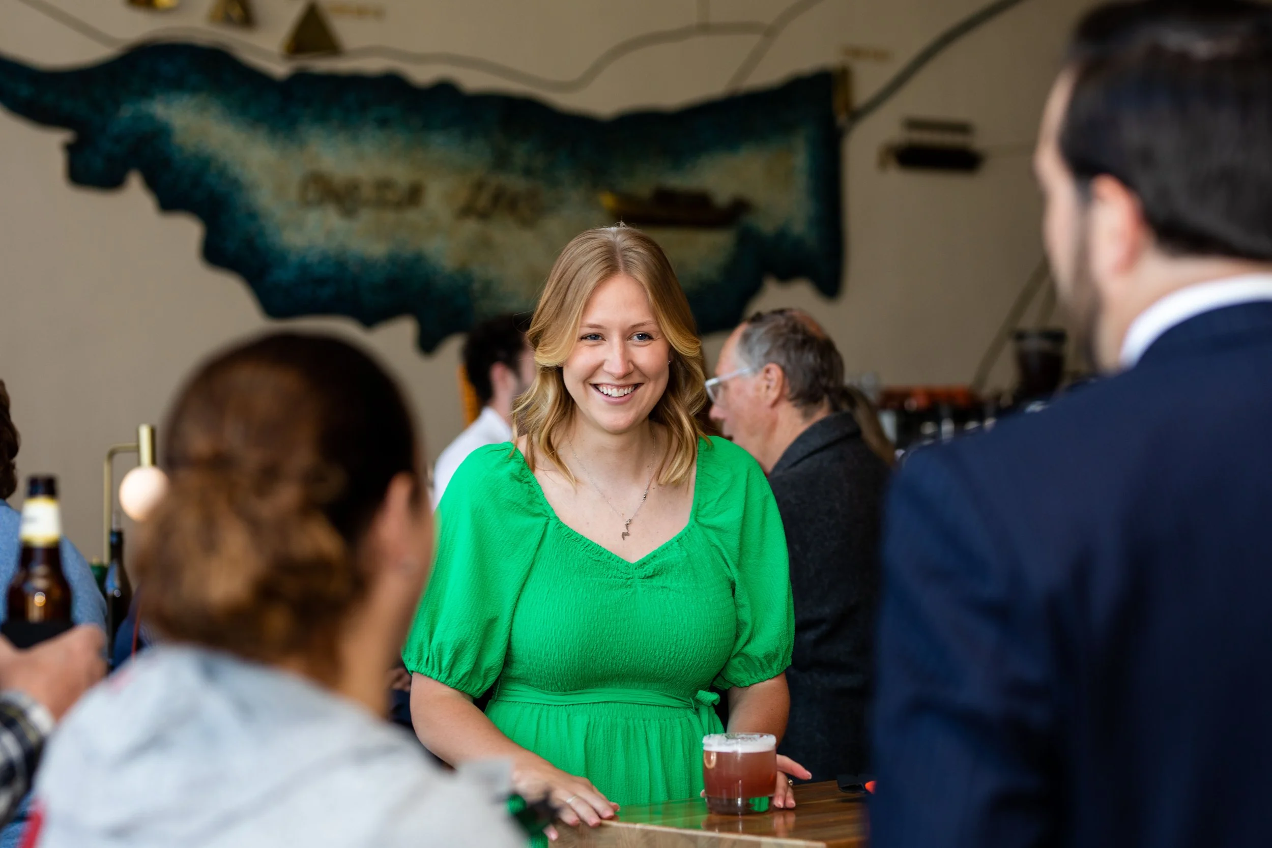 A woman in a bright green dress smiling and talking at a bar or social gathering, holding a glass of beer, with other people around and a large map of Michigan on the wall behind her.