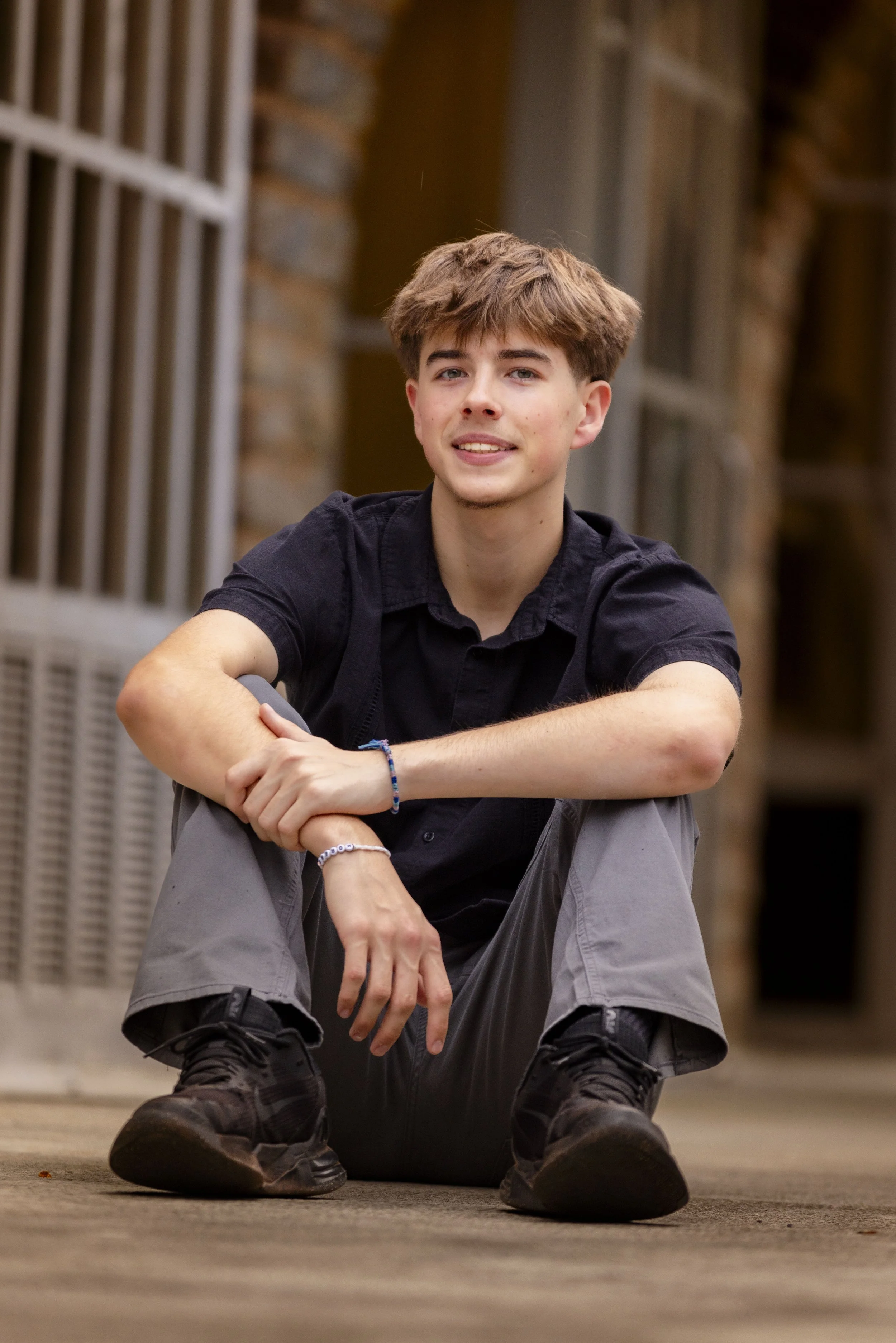 A young man with brown hair, wearing a black shirt, gray pants, and black sneakers, sitting on the ground with one knee up and the other leg crossed, smiling at the camera. Background shows brick walls and windows.