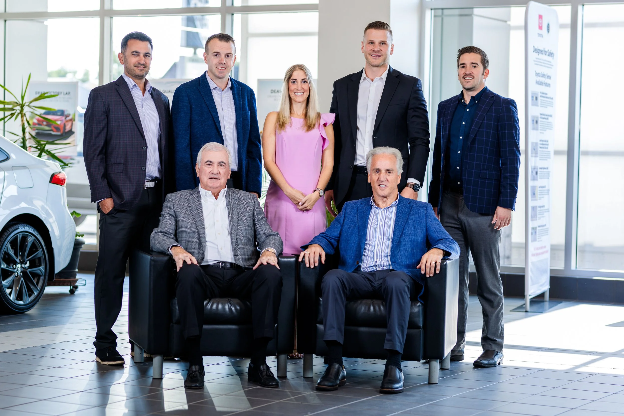 Group of nine professionally dressed people, five standing behind and two sitting on a black leather couch, in a brightly lit modern office or showroom with cars and informational posters in the background.