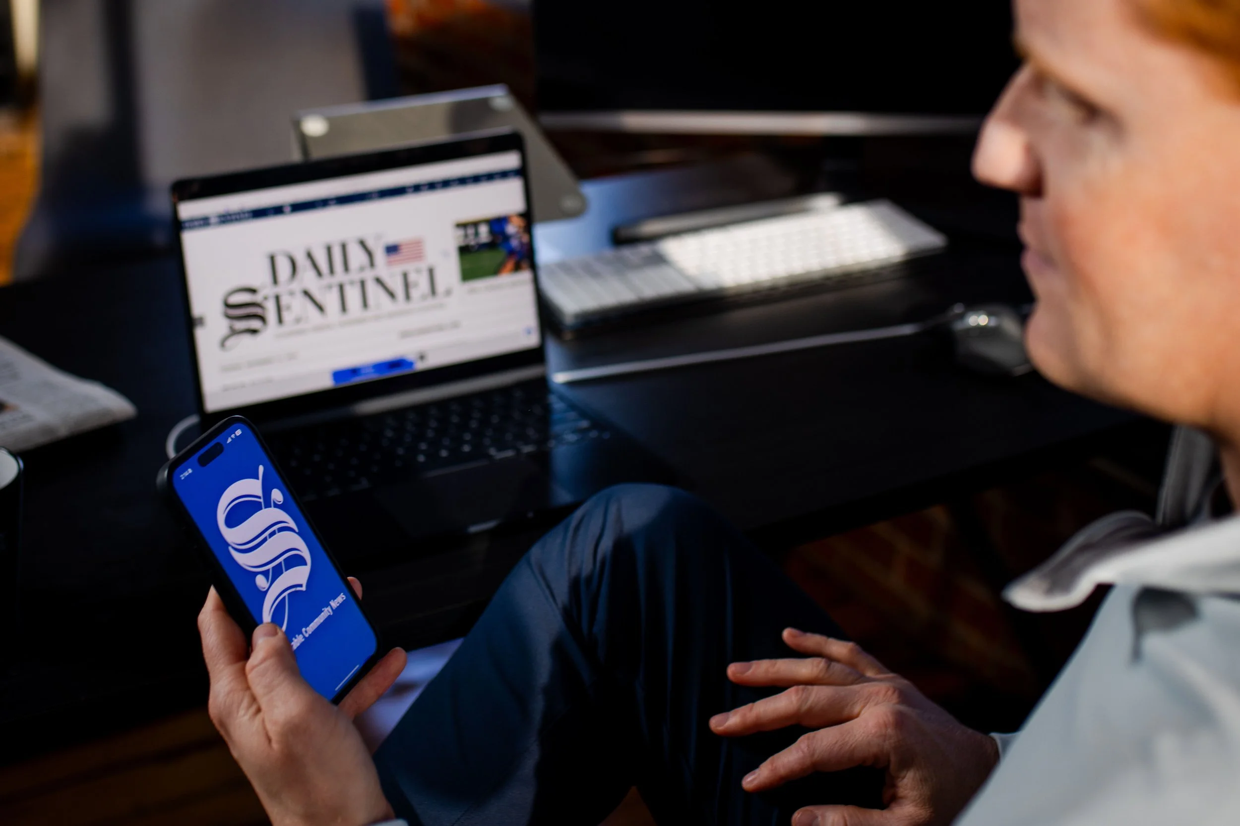 A woman sitting at a desk holding a smartphone with a logo on the screen. There is a laptop in front of her displaying a newspaper website, and a keyboard, mouse, and some papers are also on the desk.
