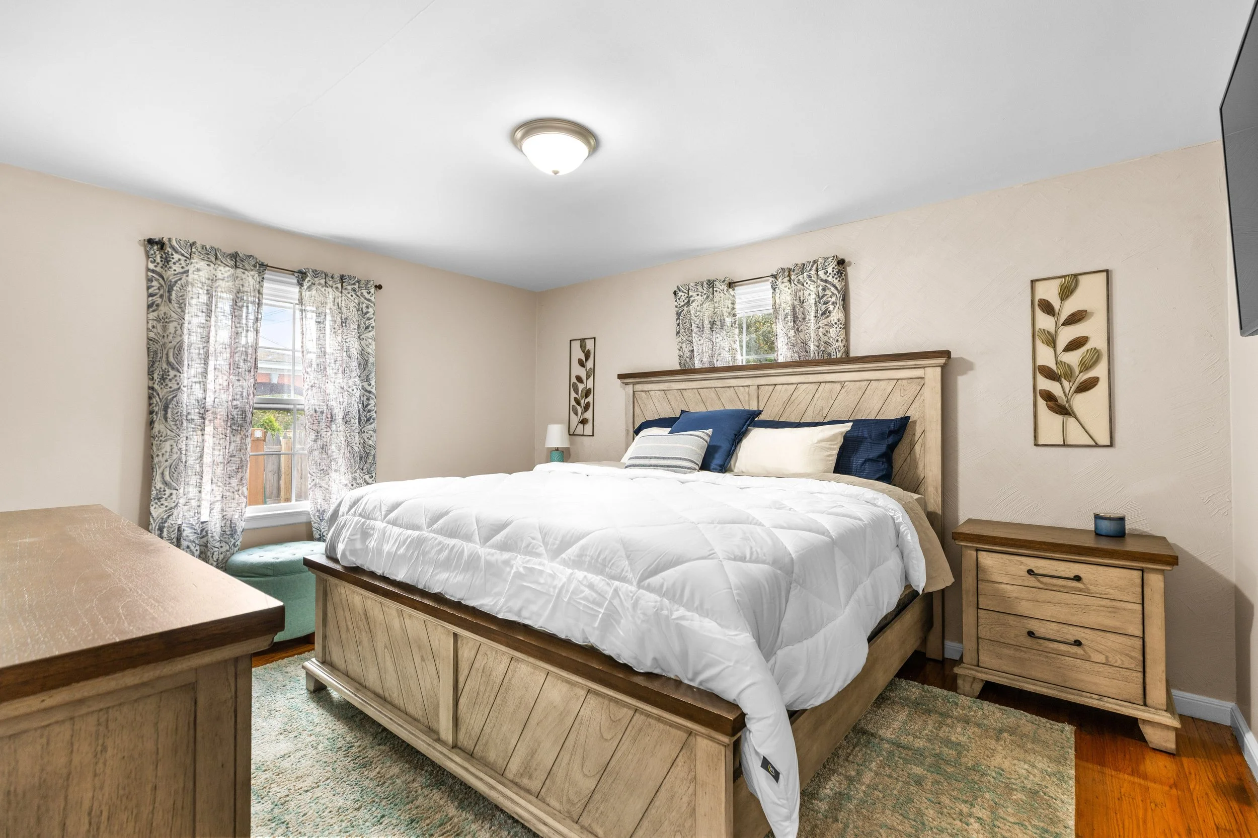 A cozy bedroom with a large wooden bed, white comforter, blue and white pillows, two windows with patterned curtains, a wooden nightstand with a candle, and decorative framed leaf artwork on the walls.