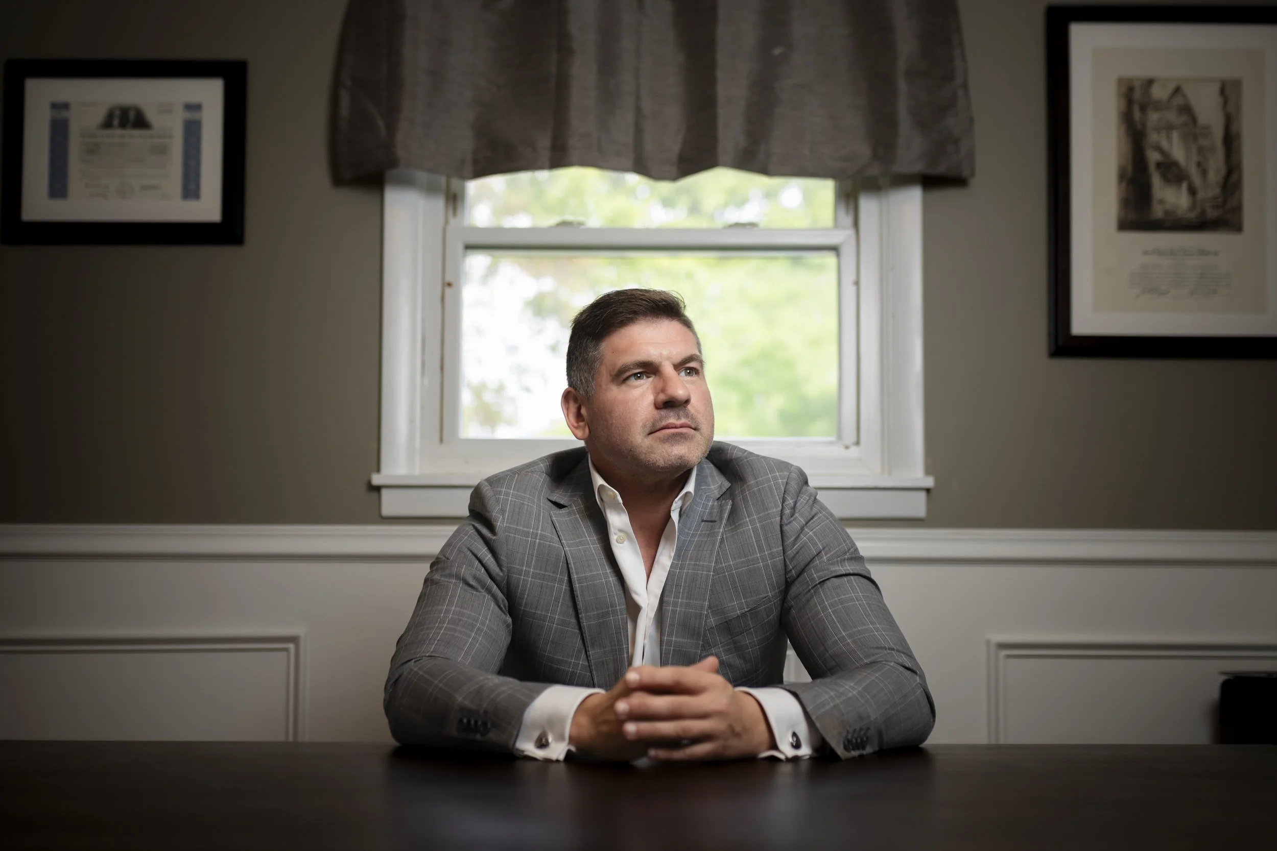 A man in a gray checkered suit sitting at a dark wooden table in a room with framed pictures on the wall and a window with a gray valance behind him.