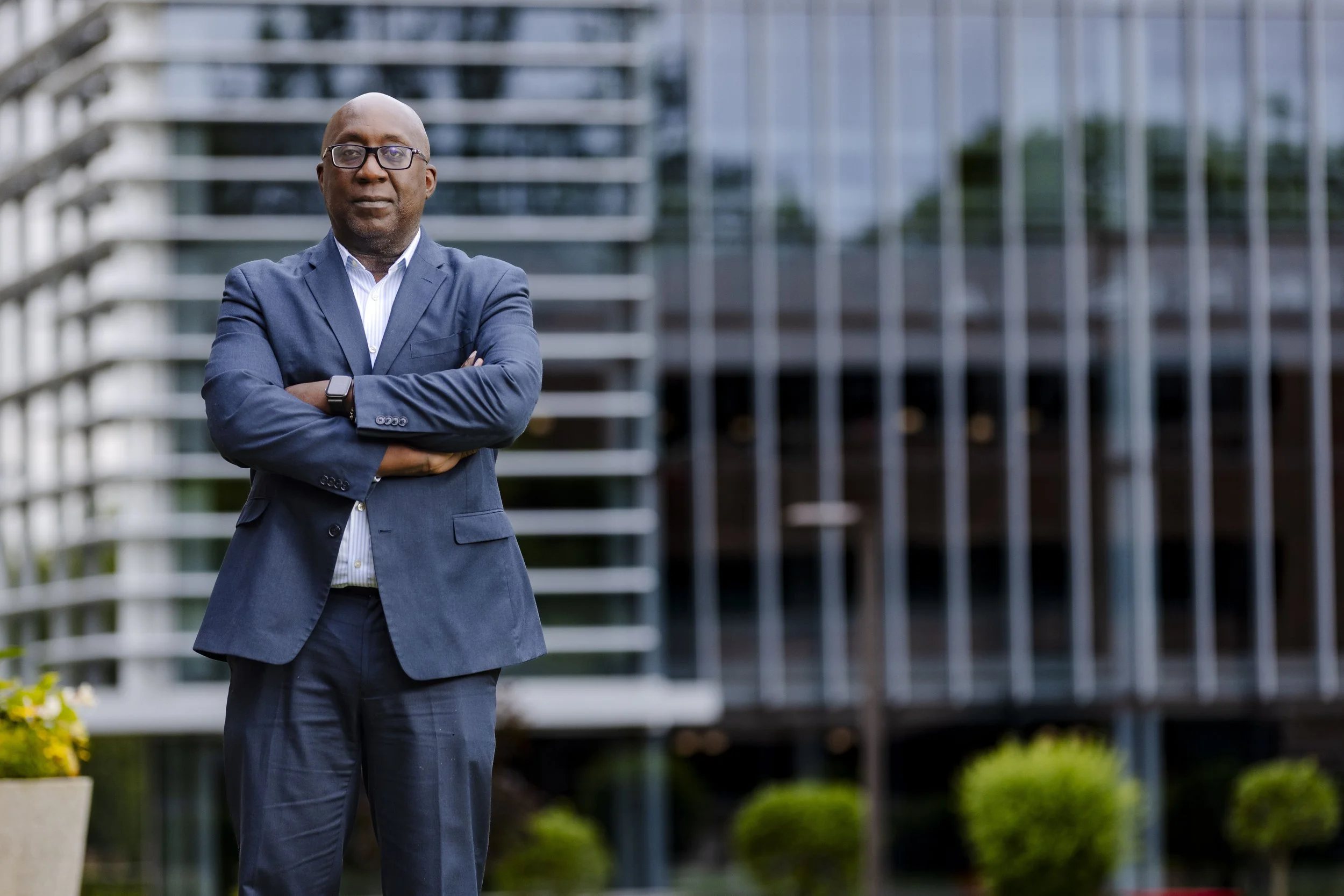 A man in a blue suit standing with arms crossed outside a modern glass building.